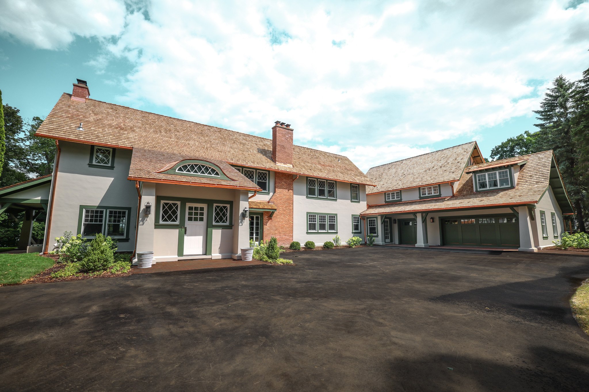 A large, two-story house with a variety of window shapes, brick chimney, and a new asphalt driveway in front, surrounded by green trees and a partly cloudy sky.