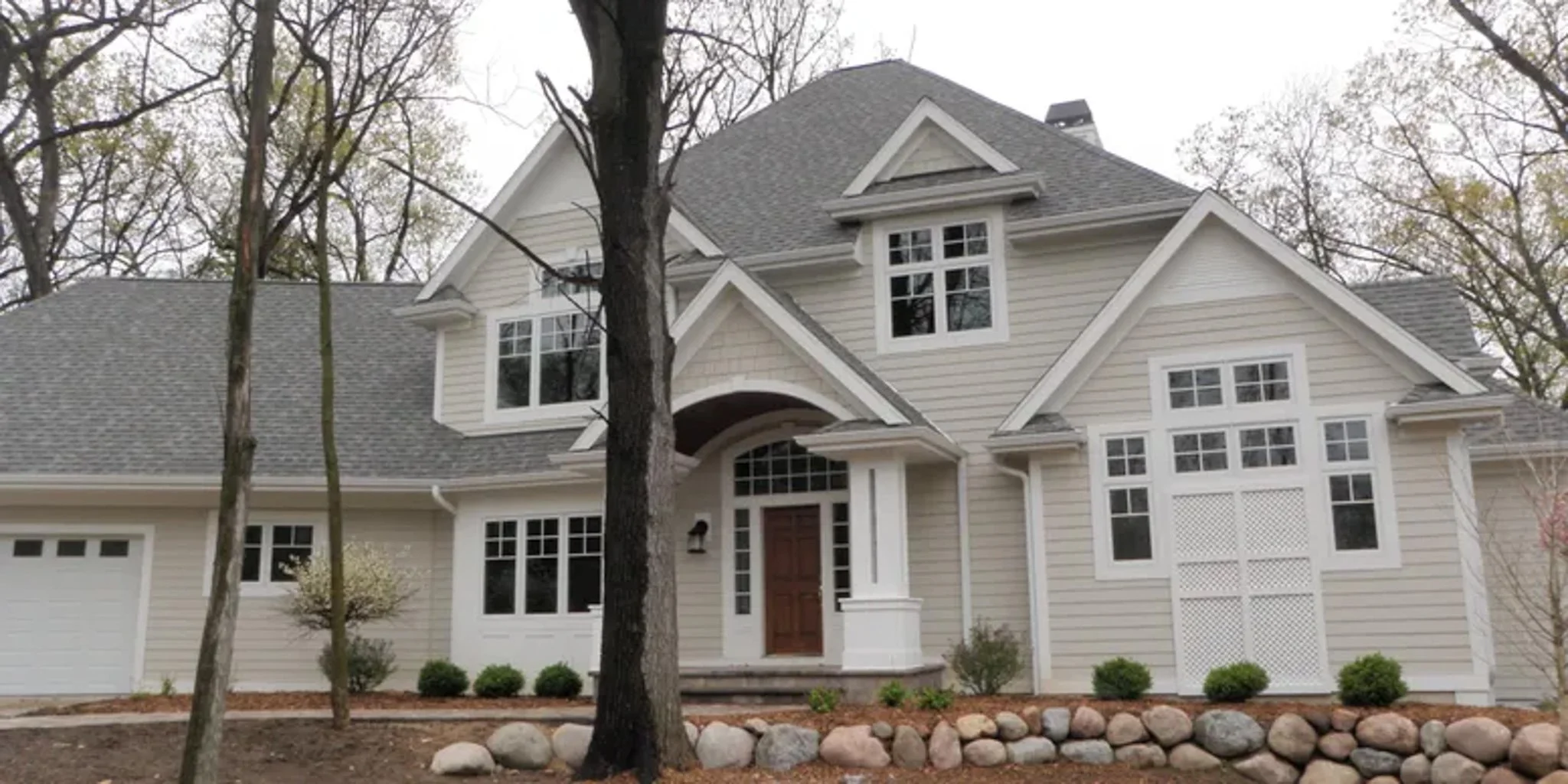 A two-story, beige house with a gray roof, multiple windows, a front door, and a garage, surrounded by trees and a landscaped yard with rocks and shrubs.
