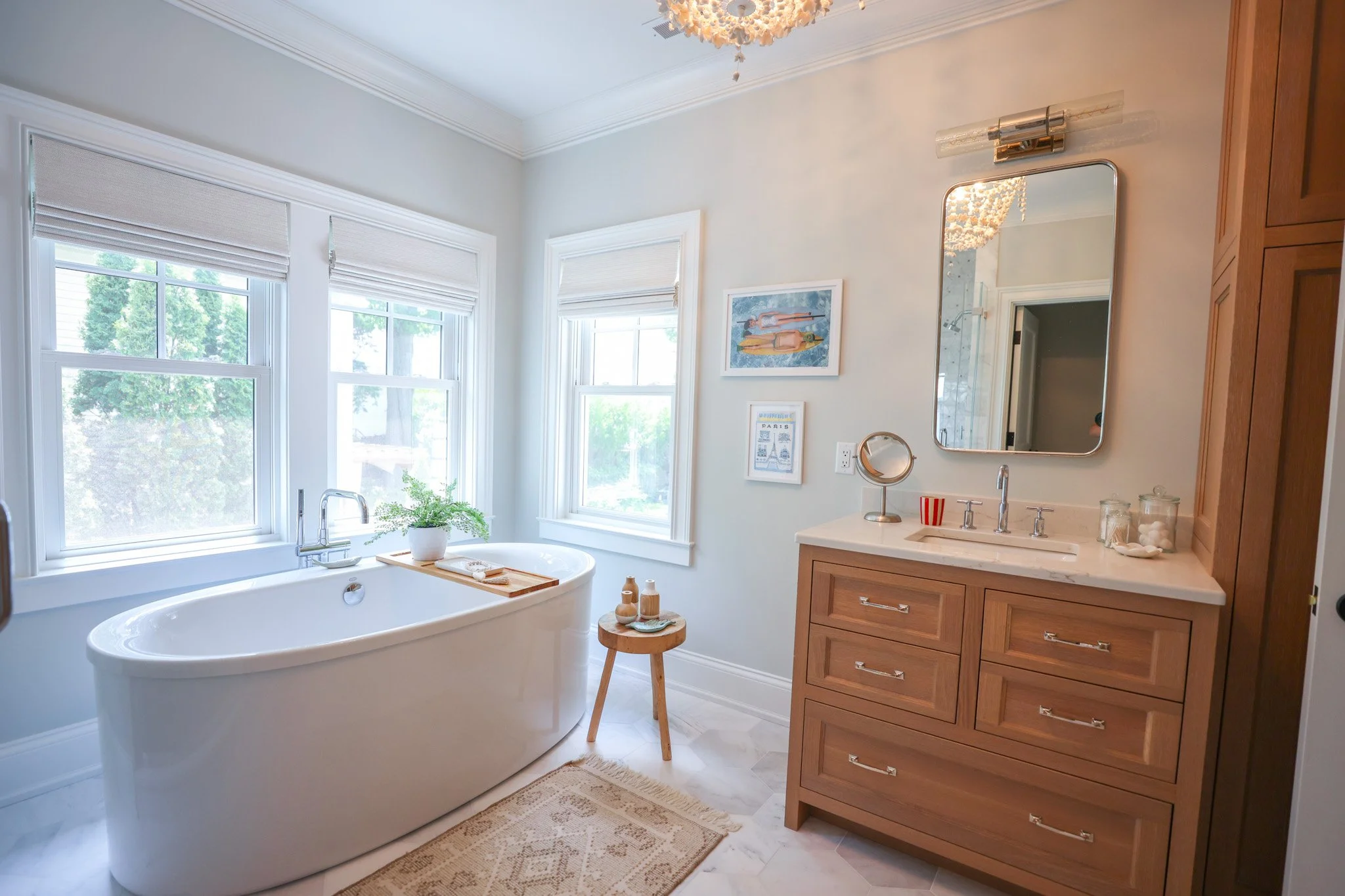 A bright bathroom with a white bathtub beneath three large windows with white blinds, a wooden stool with decorative bottles, framed artwork on the wall, a marble countertop with a faucet, and wooden cabinetry, illuminated by natural light and a ceil