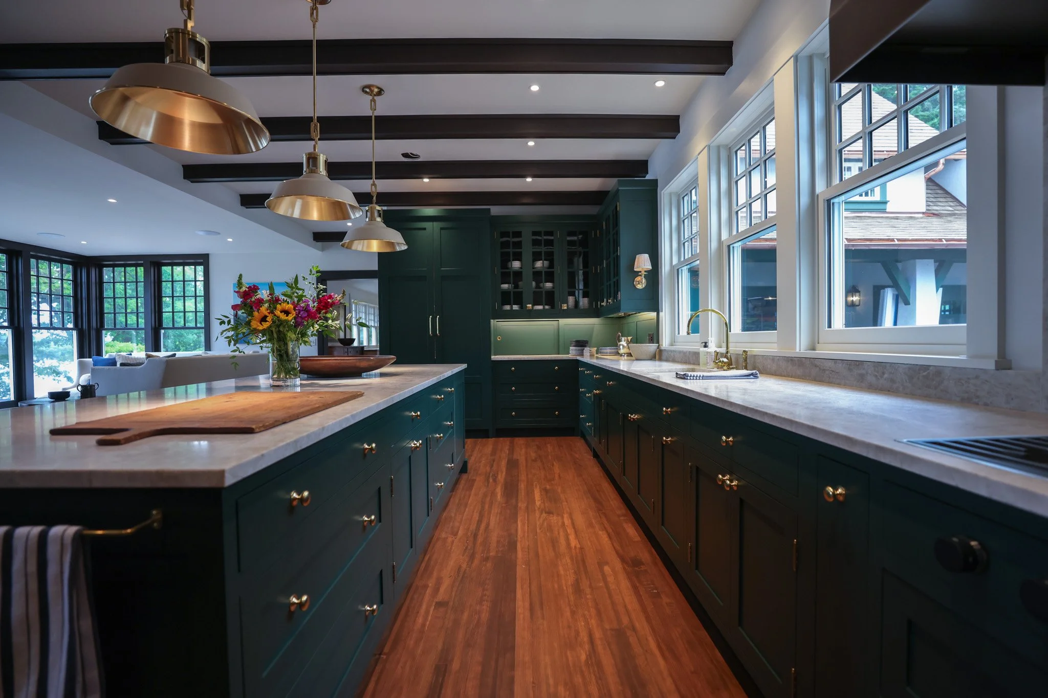 Modern kitchen with dark green cabinets, white marble countertops, large windows, pendant lights, and a flower arrangement on the island.