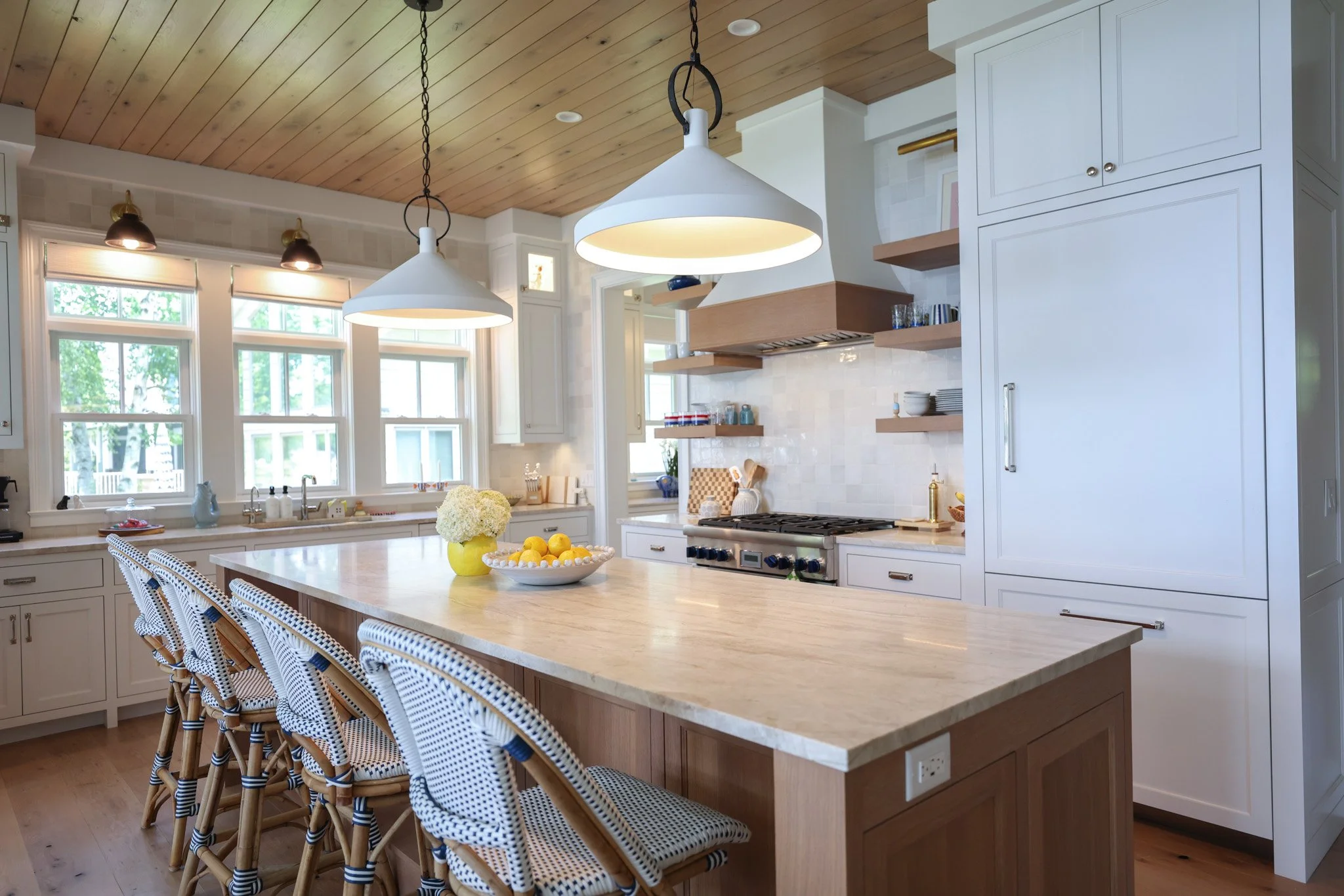A bright kitchen with a large island, white cabinets, and open wooden shelves. Two large white pendant lights hang above the island. The kitchen has wood-paneled ceiling and windows with a view of greenery outside. The island has a bowl of lemons and