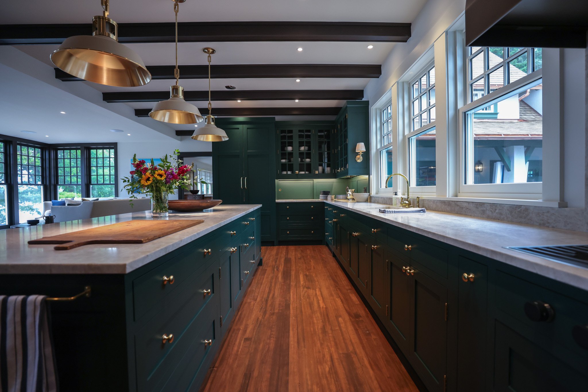 A modern kitchen with green cabinets, white marble countertops, and large windows. There are pendant lights hanging from a dark wooden beam ceiling. A vase of colorful flowers is on the island.