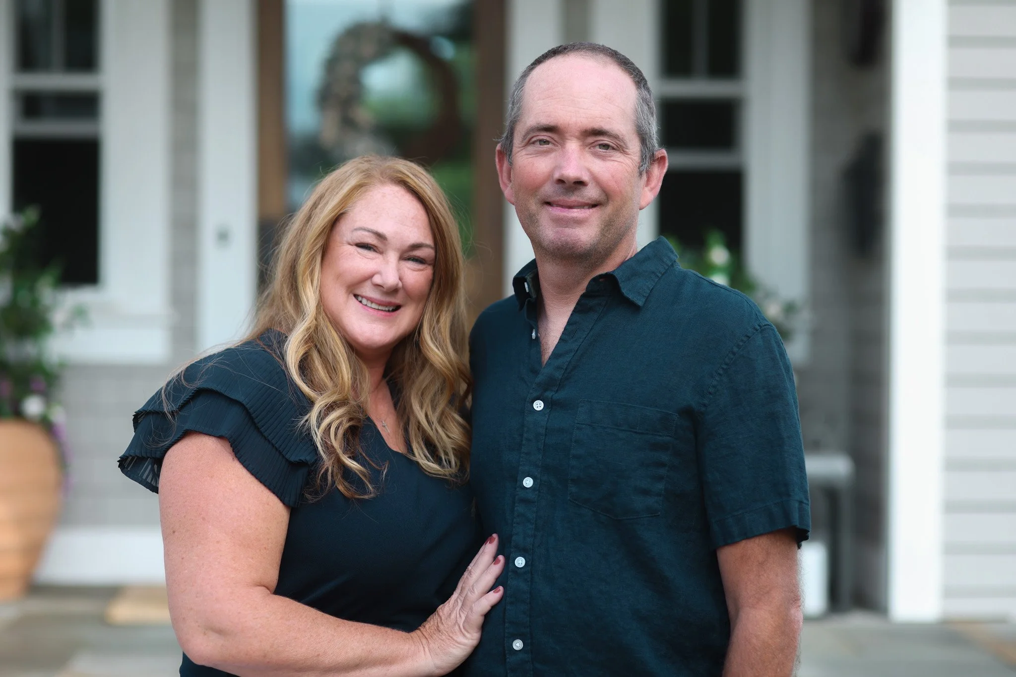 A middle-aged woman with long, wavy blonde hair and a middle-aged man with short gray hair, smiling and standing close together in front of a house with white siding and a porch. The woman has her hand on the man's chest.