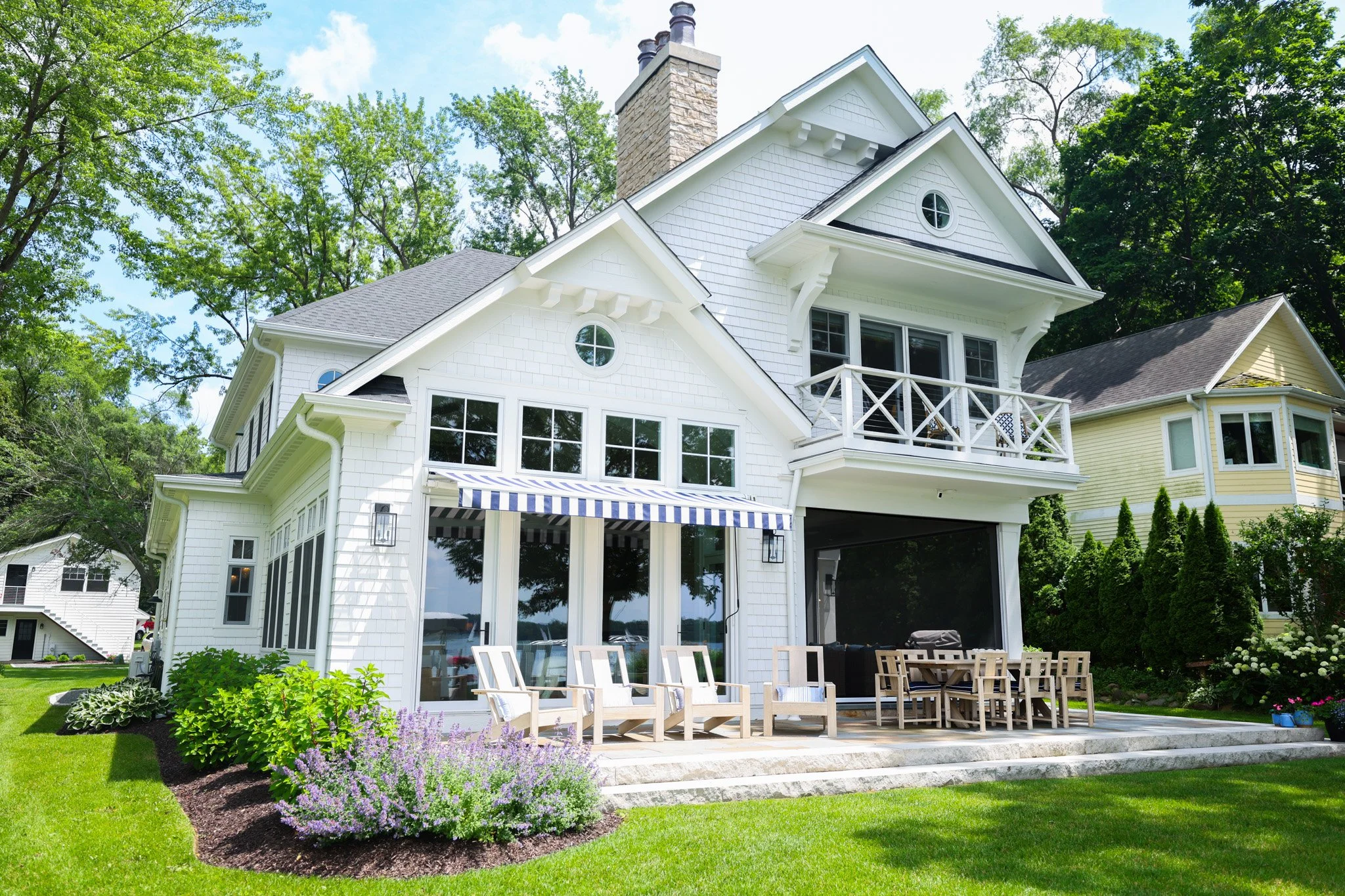 A large white house with a backyard patio, surrounded by green trees and lawn, with outdoor dining furniture and colorful flowers.