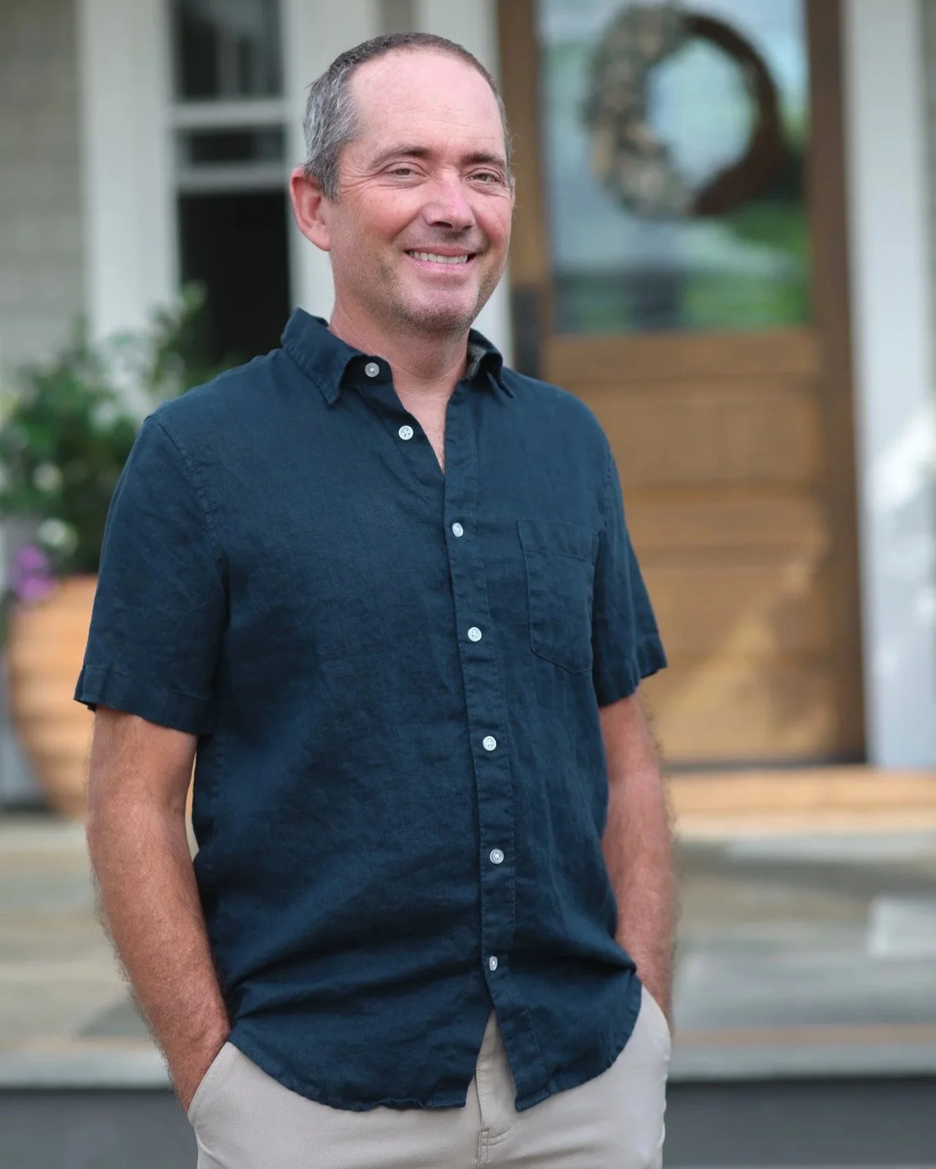 A man standing outdoors in front of a house, smiling, wearing a dark blue button-up shirt and beige pants.