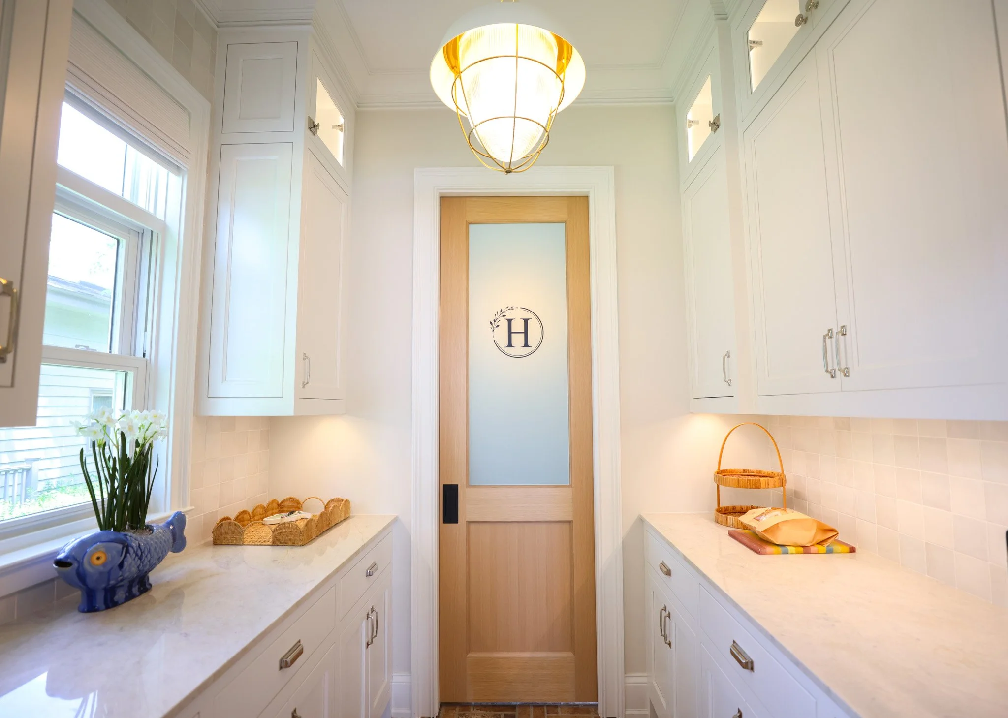 A bright kitchen with white cabinetry, a beige marble countertop, and a wooden door with a frosted glass panel featuring the letter 'H' inside a circle. There are windows on both sides, sunlight coming in, and decorative items like a blue fish-shaped