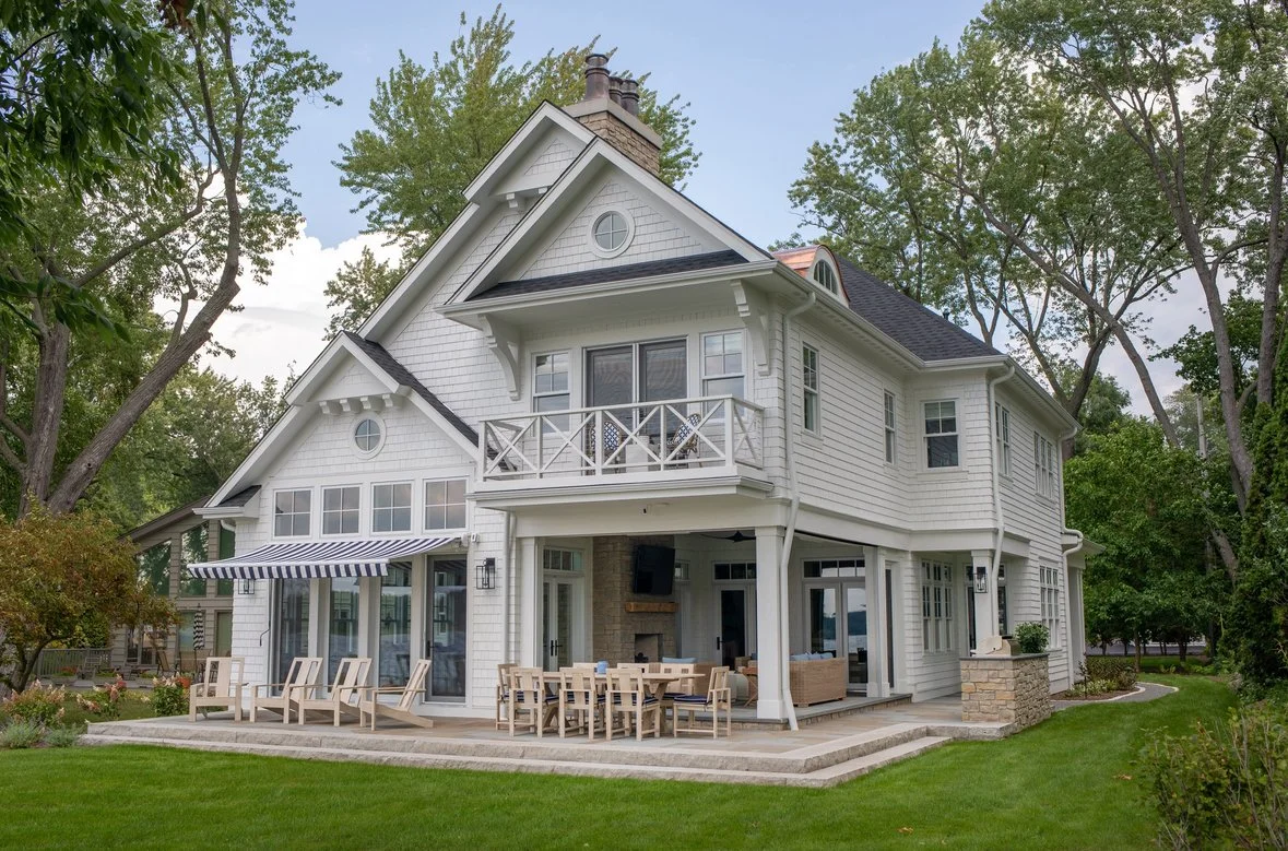 A large white two-story house with a patio and outdoor dining area, surrounded by green trees and lawn.