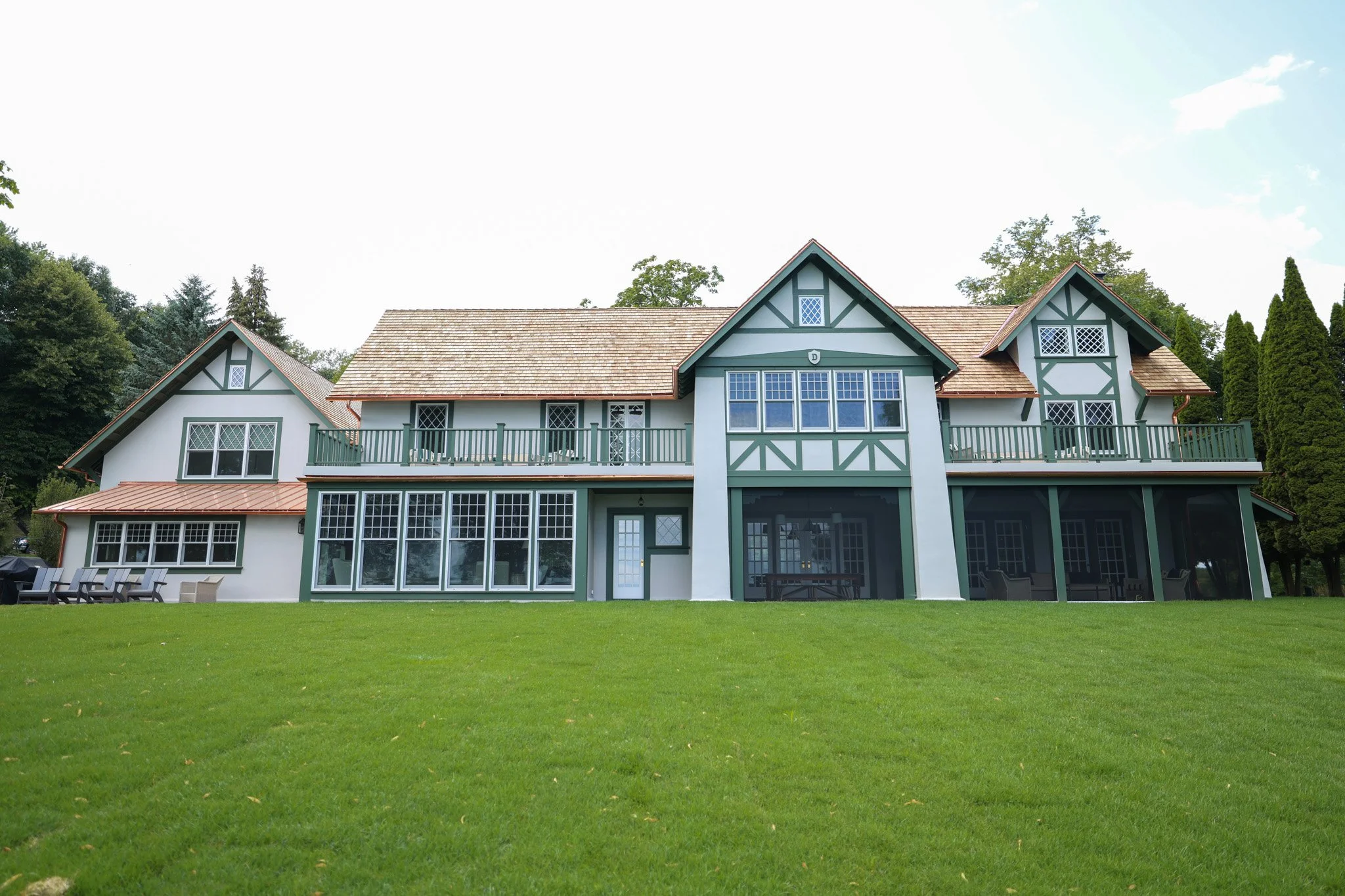 A large, house with green and white exterior, multiple roof peaks, and a wrap-around balcony, surrounded by a green lawn and trees.
