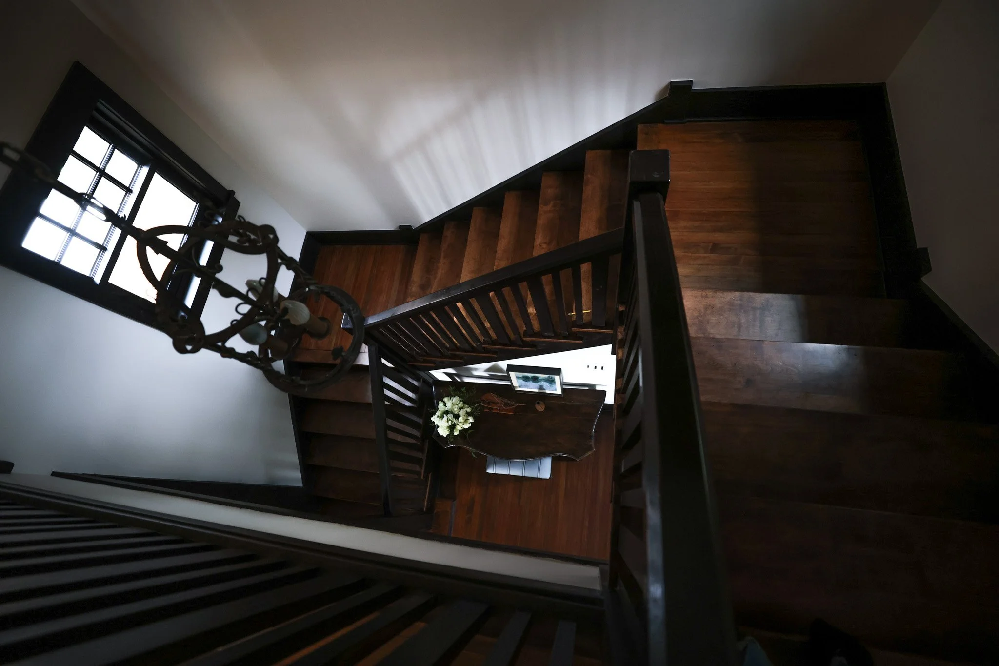 Interior view of a staircase with dark wooden steps, black railings, a chandelier, a window letting in natural light, a bookcase with a monitor, and a vase of white flowers on a table.