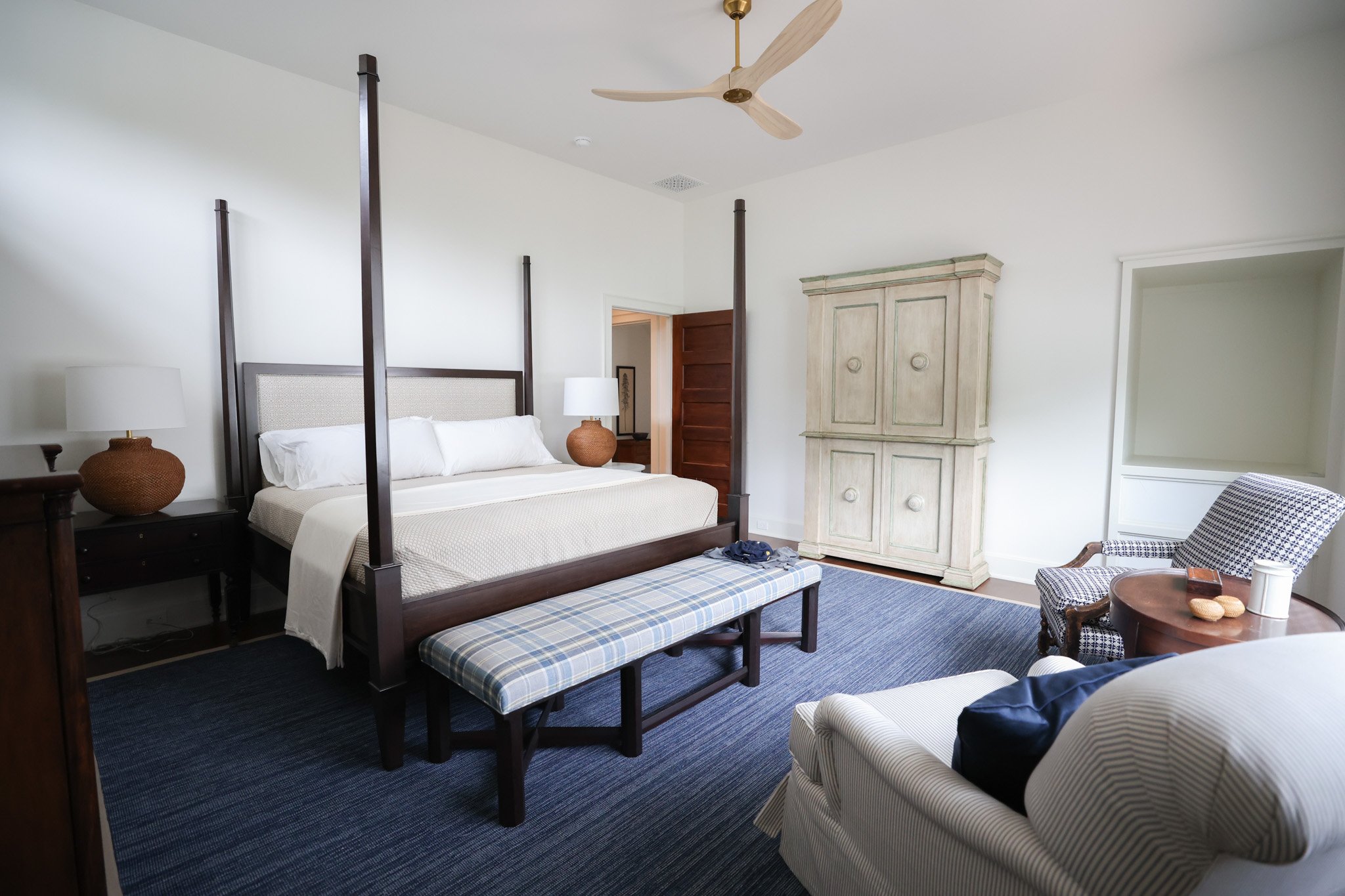 A bedroom with a four-poster bed, two nightstands with lamps, an armchair, and a wooden wardrobe, with a blue carpet and plain white walls.