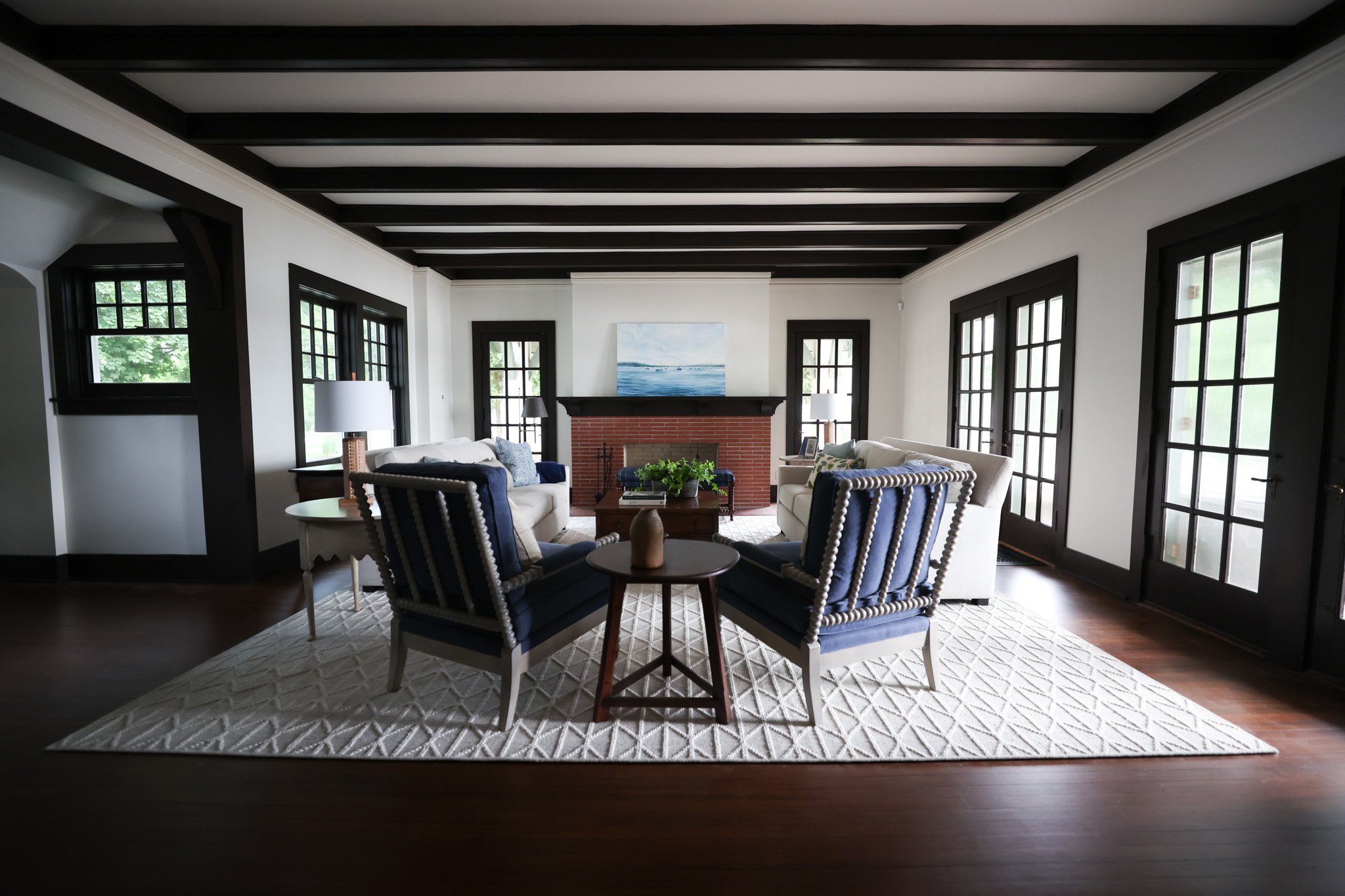 Living room with white walls, dark wooden ceiling beams, large windows with black frames, a brick fireplace, and beige and blue furniture, including sofas and chairs, arranged around a wooden coffee table with a potted plant, a painting of water abov