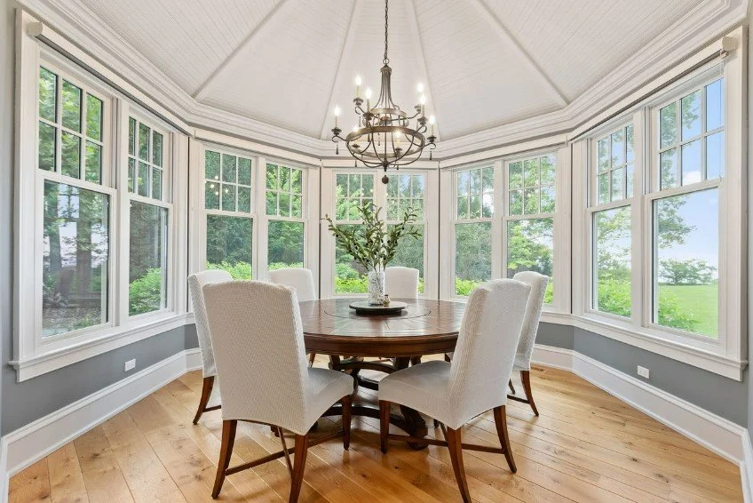 A bright dining room with large windows, a round wooden table, six upholstered chairs, a chandelier, and a vase with greenery, overlooking a lush outdoor scene.