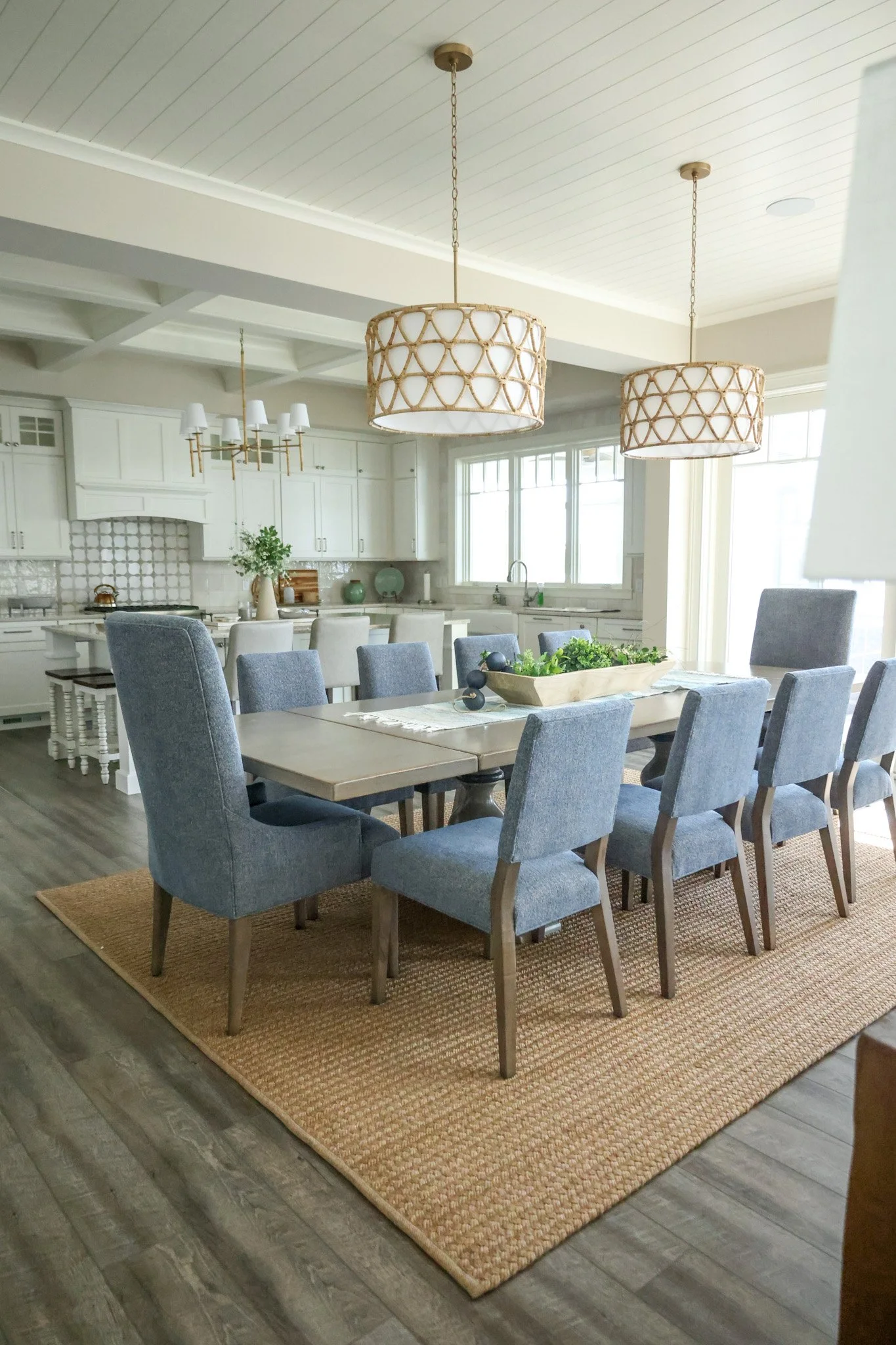 A dining area with a rectangular wooden table surrounded by blue upholstered chairs, on a brown woven rug. A rectangular wooden tray with green plants is on the table. The room has a kitchen in the background with white cabinets and a window. Two lar
