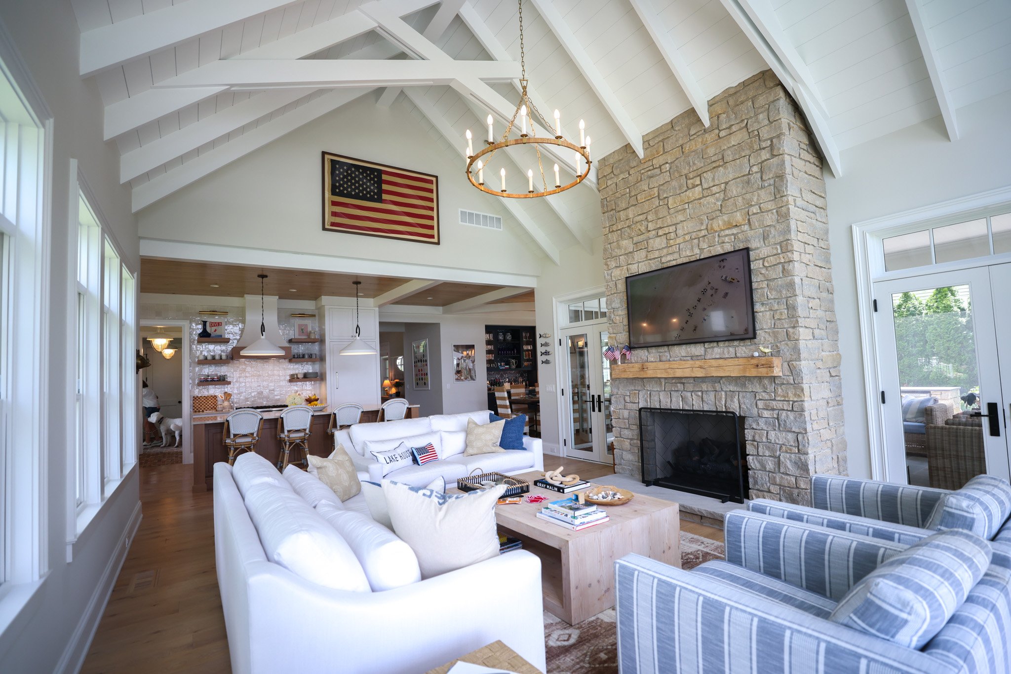 Living room with white sofas, patterned armchair, wooden coffee table, stone fireplace with a TV, American flags, and a chandelier, with large windows and an open kitchen area in the background.