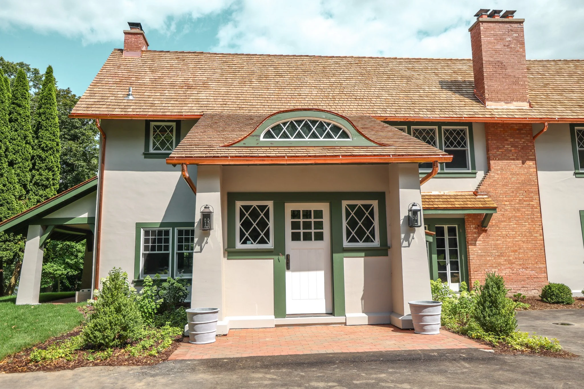 Front exterior of a modern house with a green, white, and brick facade, large front porch, and brick chimney, surrounded by a landscaped garden with bushes and trees.