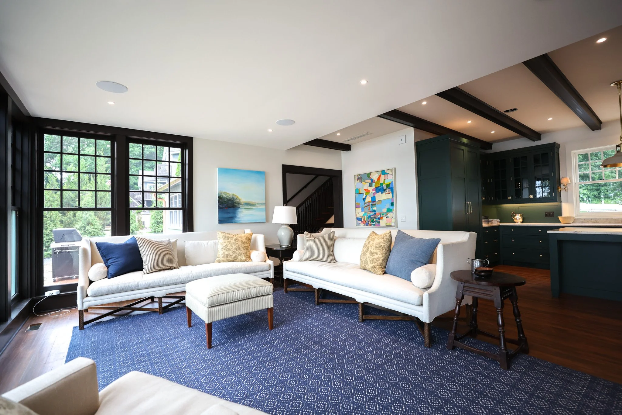 Bright living room with large black-framed windows, white sofas with patterned pillows, artwork on white walls, a dark blue cabinet, and a blue patterned rug.