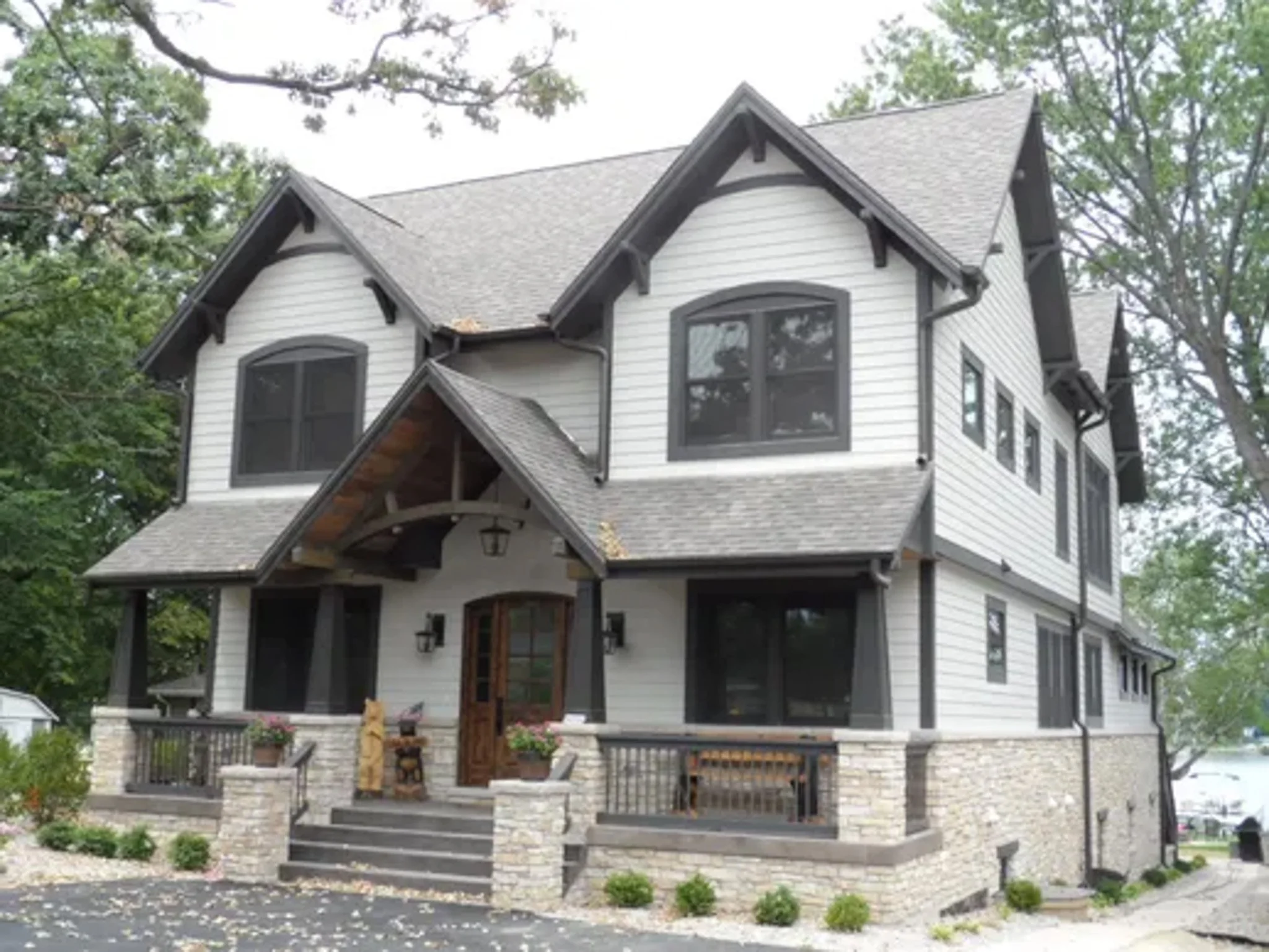 Front view of a two-story house with gray gabled roof, white siding, black window frames, and a stone foundation, surrounded by trees and a concrete walkway with small bushes.
