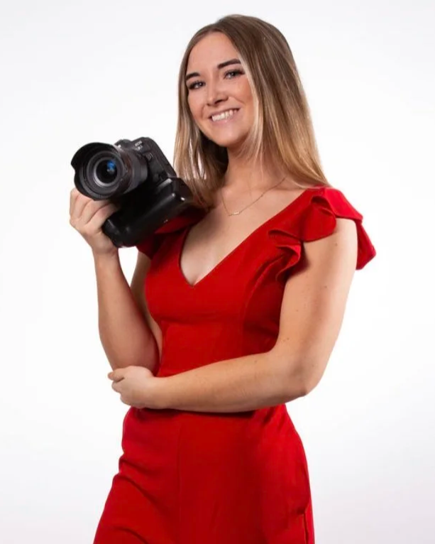 A young woman with long, light brown hair smiling and holding a professional camera, wearing a red dress with ruffled sleeves on a white background.