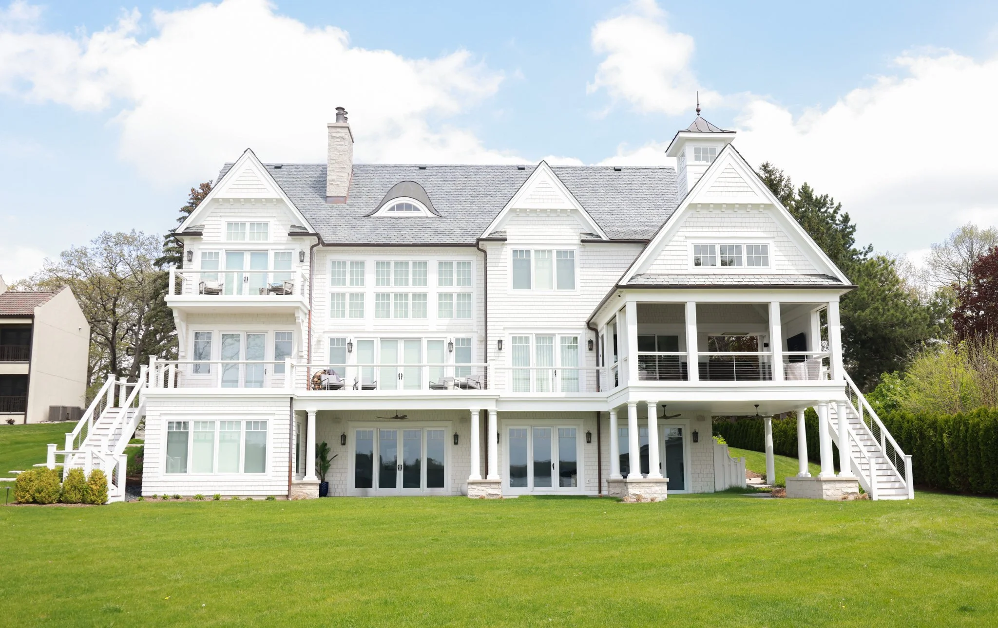 A large white multi-story house with multiple gabled roofs, large windows, and a spacious backyard with green grass and trees under a partly cloudy sky.