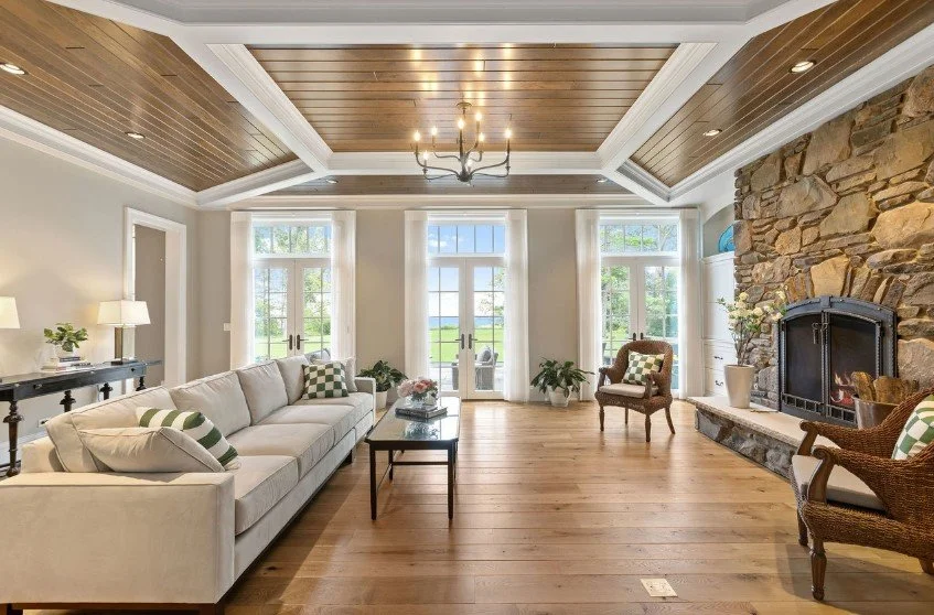 Spacious living room with large windows, stone fireplace, hardwood floors, and white walls. Features a white sofa, armchairs, and a coffee table.