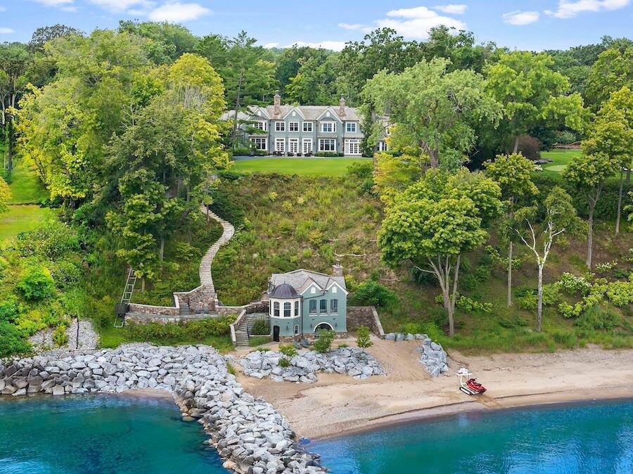 A large, elegant house on a hill overlooking a beach with colorful waters. It has a detached garage, a landscaped yard, and stairs leading down the hill. Trees surround the property, creating a lush, green environment.