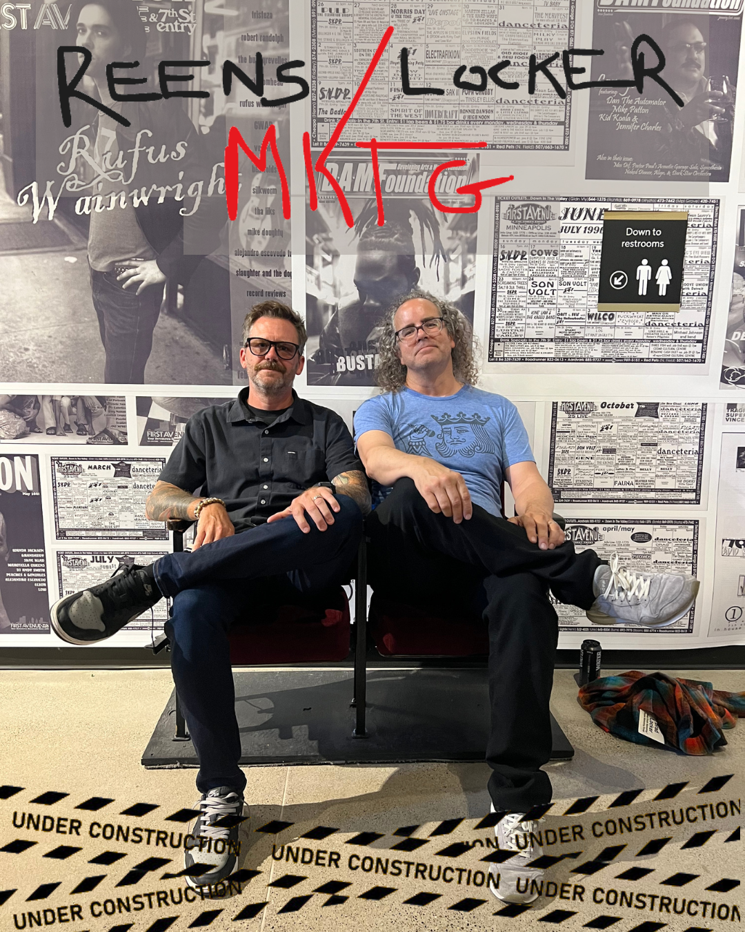 Two men sitting on a red bench in front of a black-and-white collage of posters and photos on a wall. The man on the left has glasses, a beard, and tattoos, wearing a black shirt. The man on the right has curly hair, glasses, and is wearing a blue t-shirt with a graphic design. There is a sign indicating the restrooms behind them. The floor is marked with "Under Construction" tape. The background includes various posters, a large handwritten sign, and some scattered items on the ground.