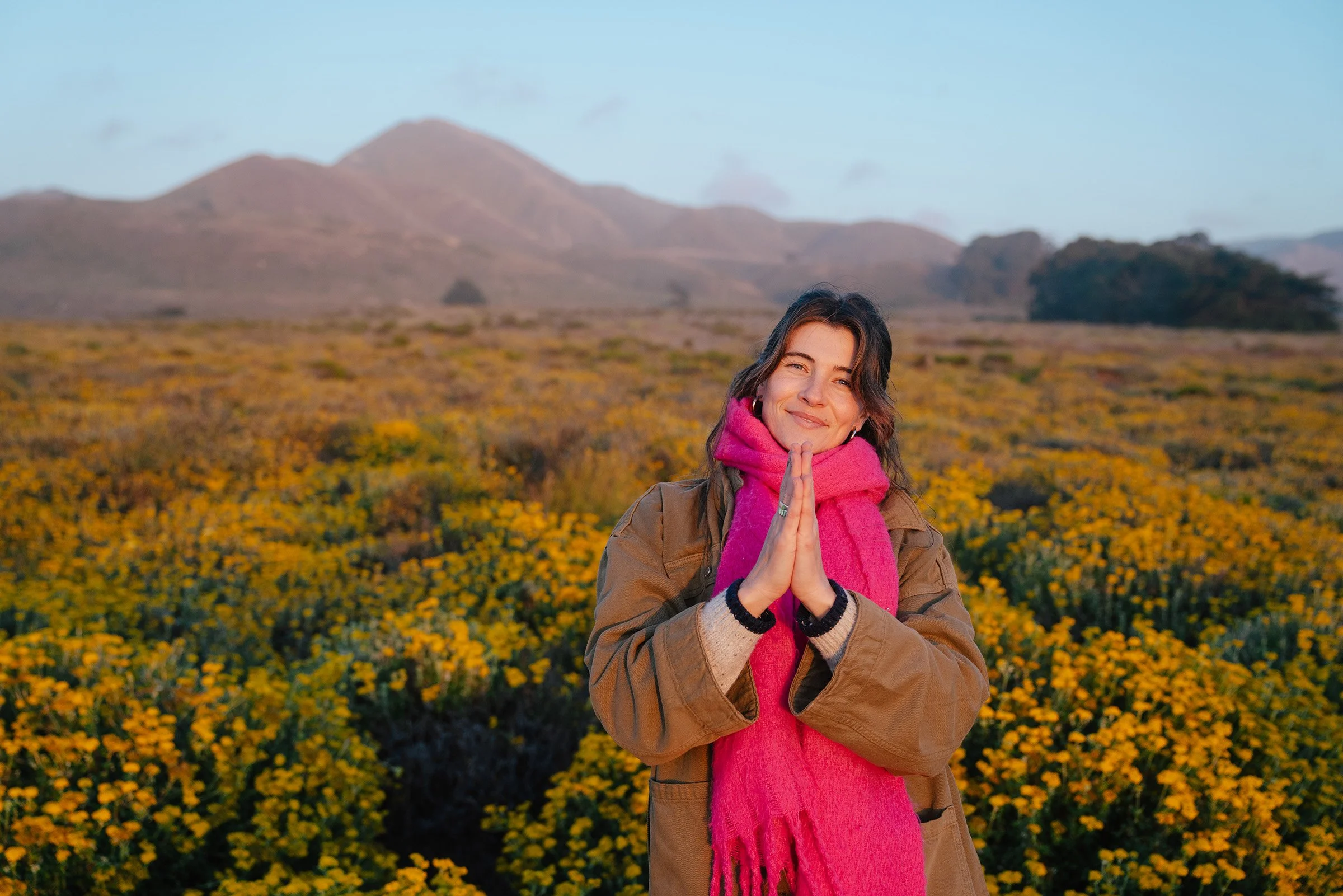 A woman standing in a field of yellow flowers, performing a prayer pose with hands pressed together, smiling, with mountains in the background.