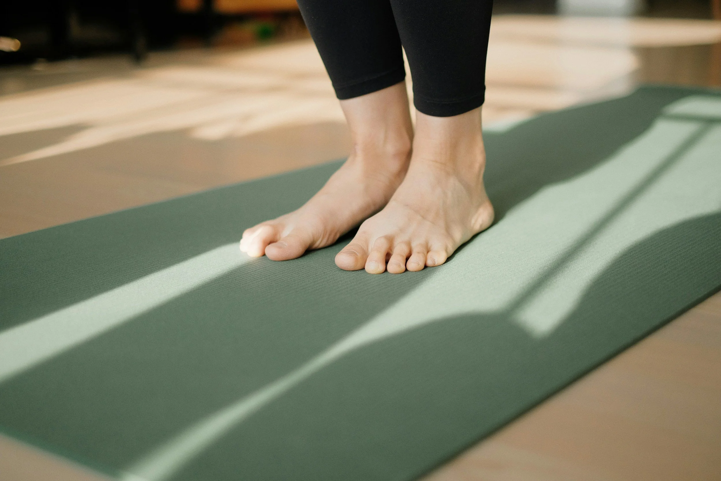A person practicing yoga or stretching on a green exercise mat, with their hands on the mat and feet slightly apart, in a room with a wooden floor.
