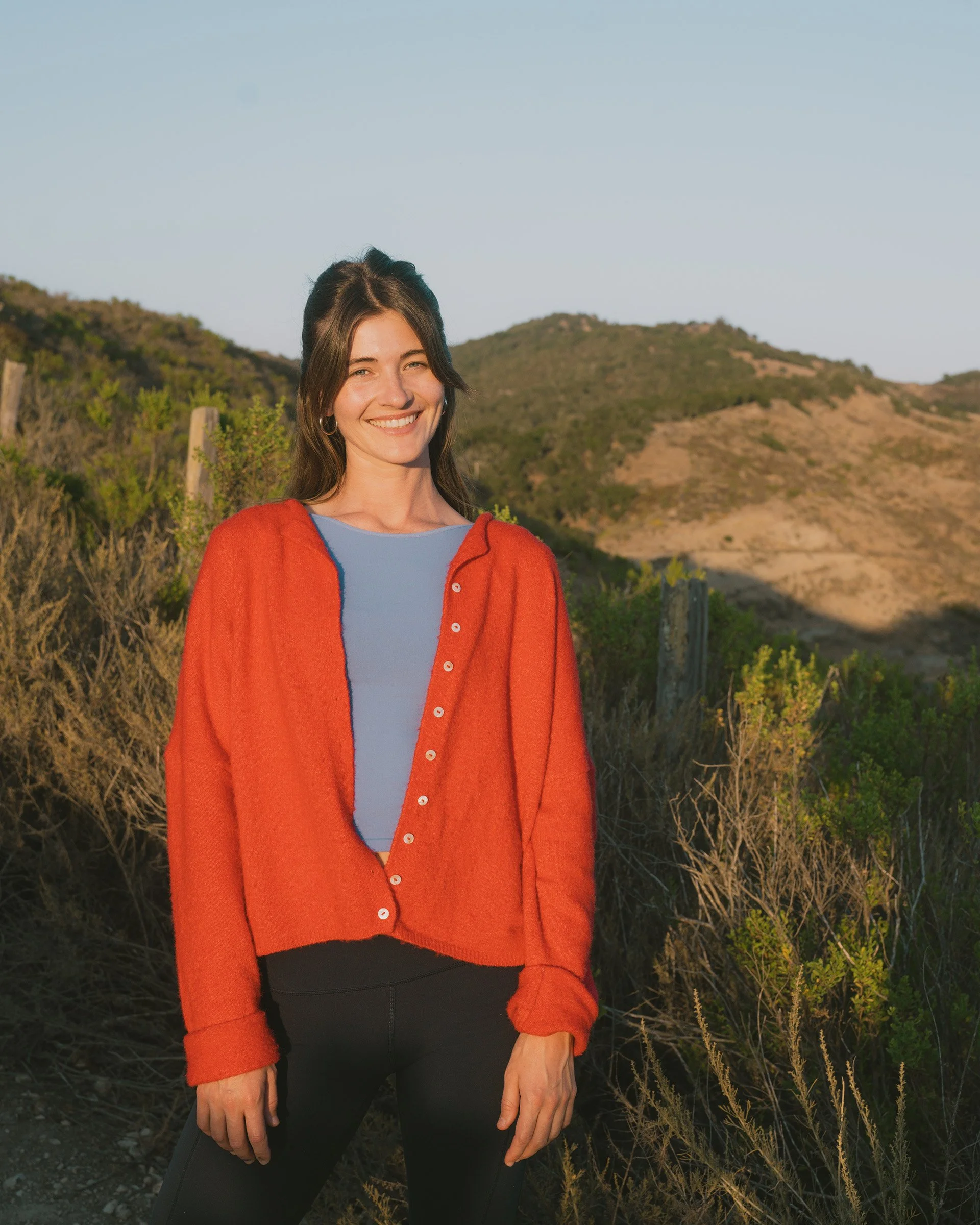 A young woman in a red sweater over a light blue top, standing outdoors in a hilly, sunny landscape with bushes and dry grass, smiling at the camera.