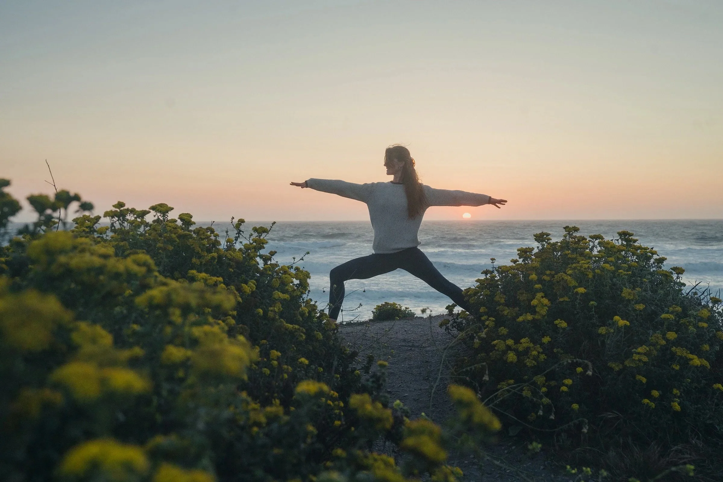 A person practicing yoga in a Warrior pose on a hill overlooking the ocean at sunset, surrounded by yellow flowering plants.