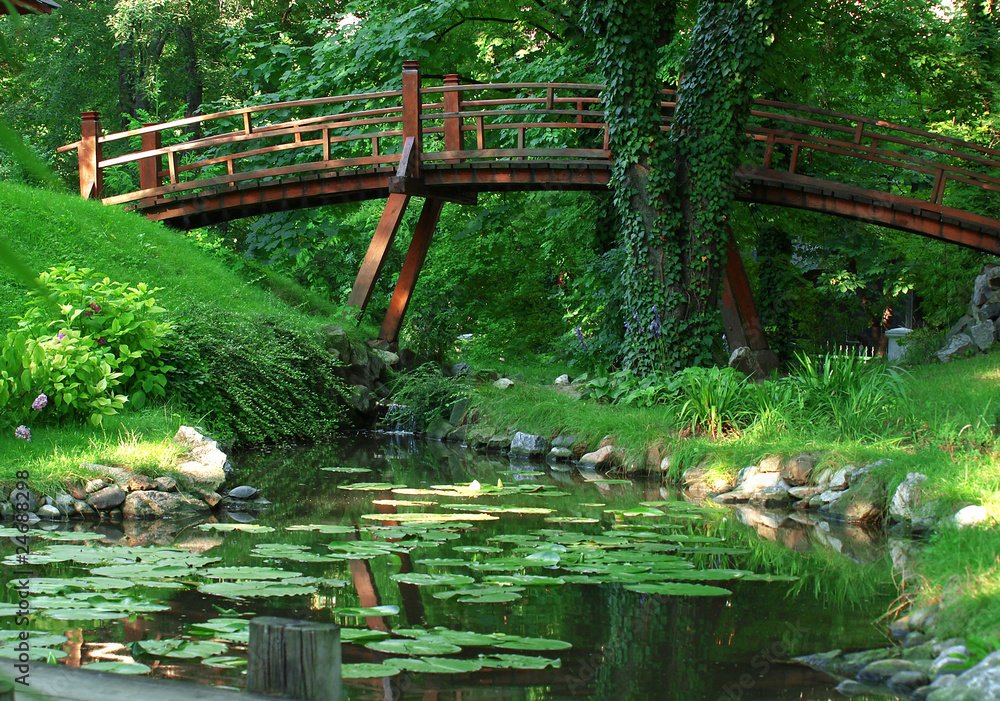 A red wooden bridge arches over a small pond with lily pads in a lush green park.