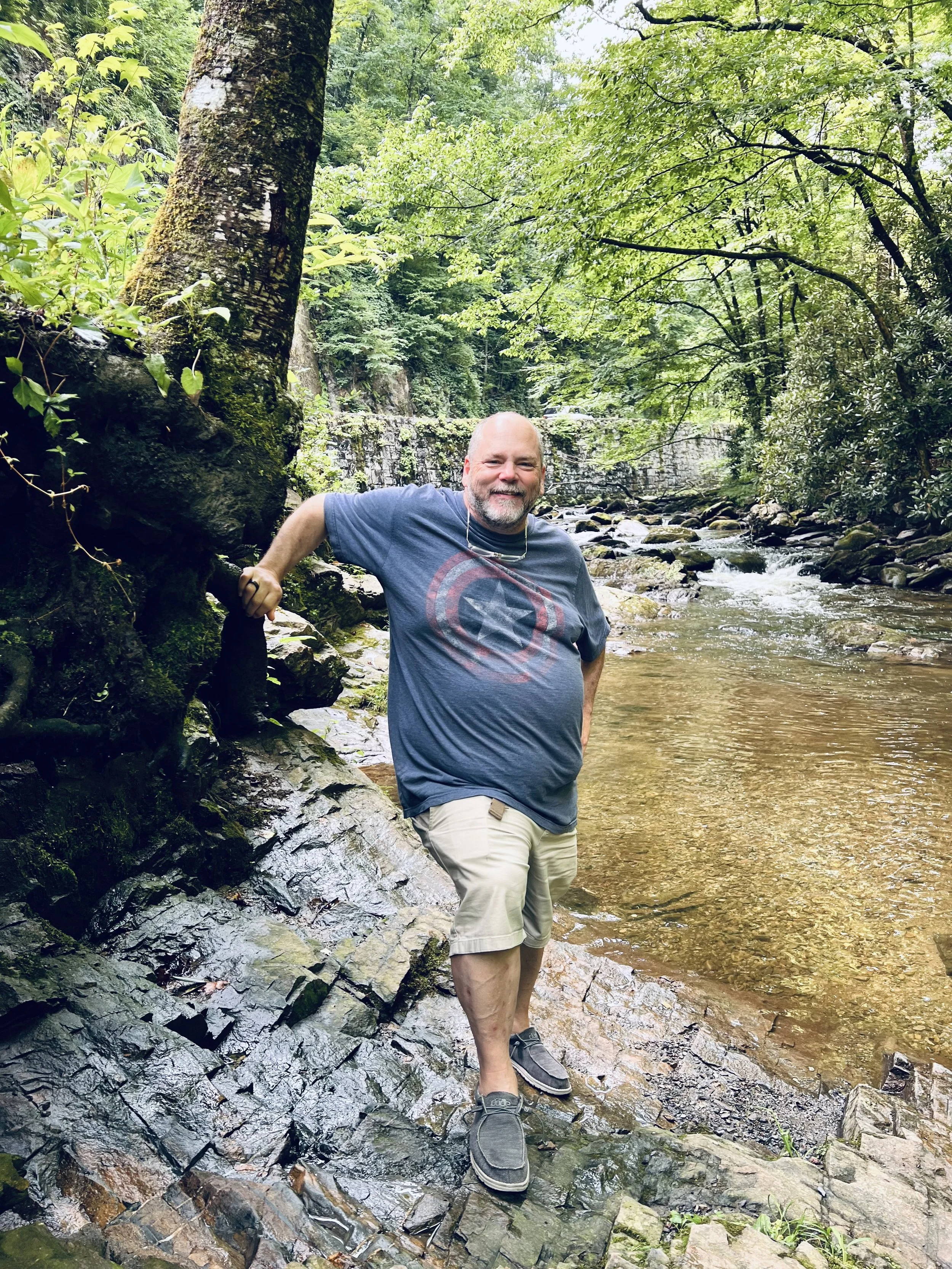 A man with a bald head and a gray beard smiling while standing on rocks in a shallow creek surrounded by lush green trees and foliage.