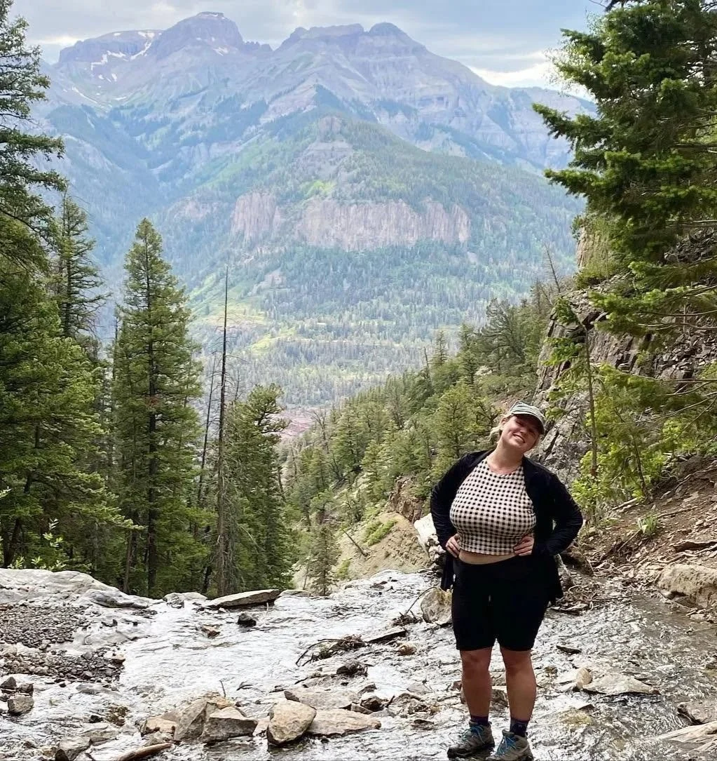 A woman standing in a rocky mountain stream surrounded by tall evergreen trees with a mountain range in the background.