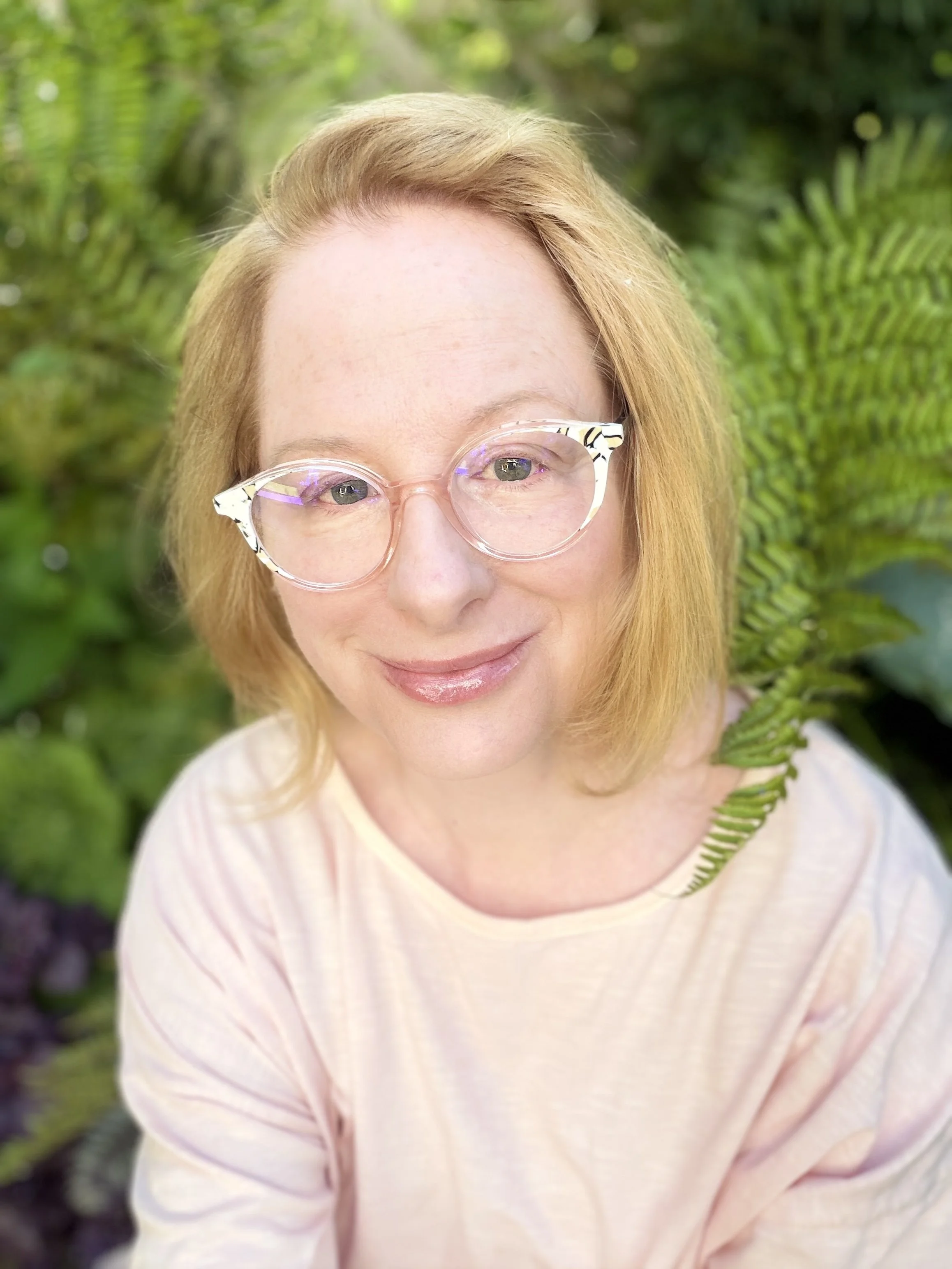 A woman with ginger hair and glasses smiling outdoors among green plants.