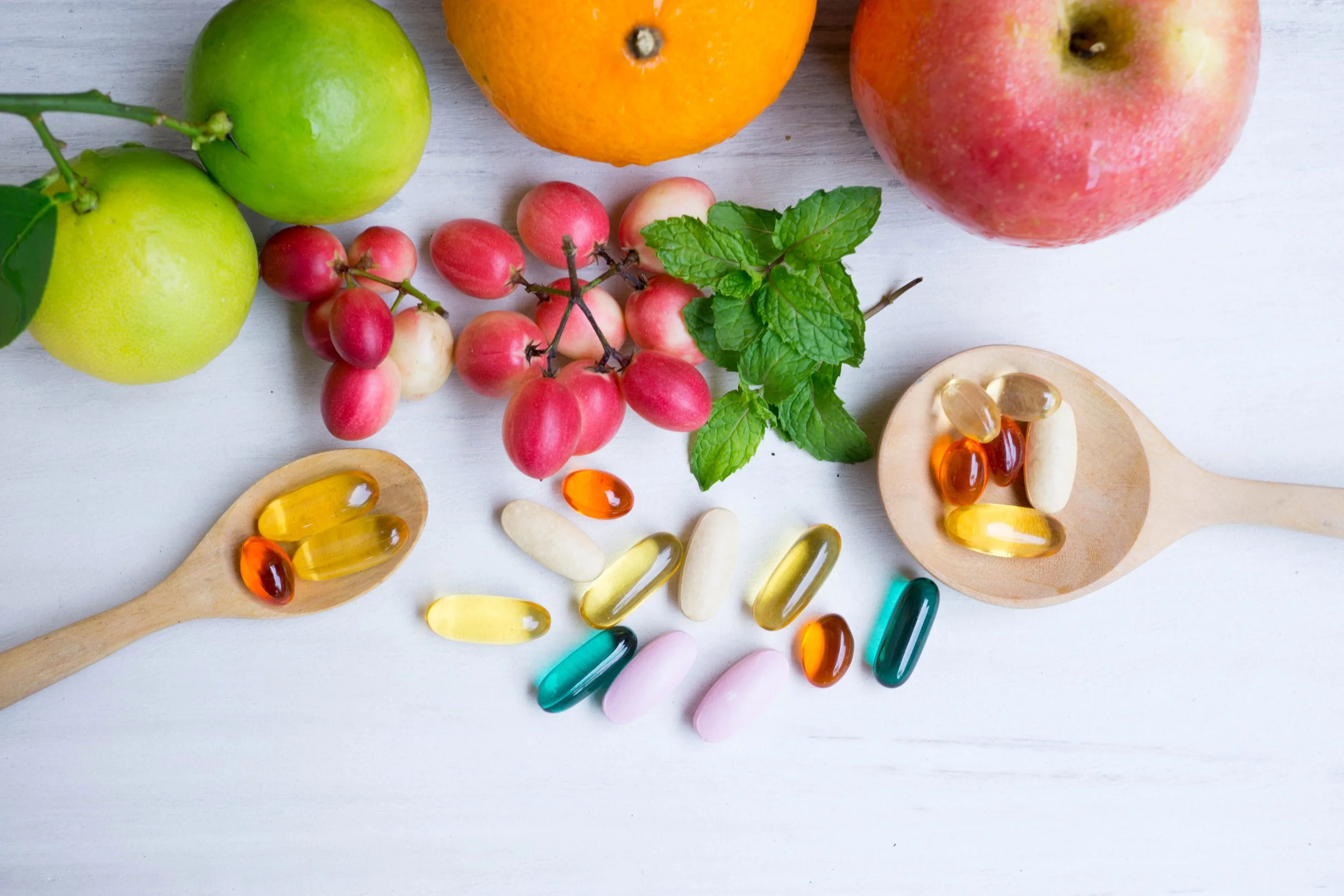 A variety of fresh fruits including green apples, a lemon, pink and white grapes, an orange, and a red apple, along with herbal mint leaves, are arranged on a light-colored wooden surface. There are also two wooden spoons filled with various capsules and pills in different colors like yellow, orange, white, pink, green, and black.