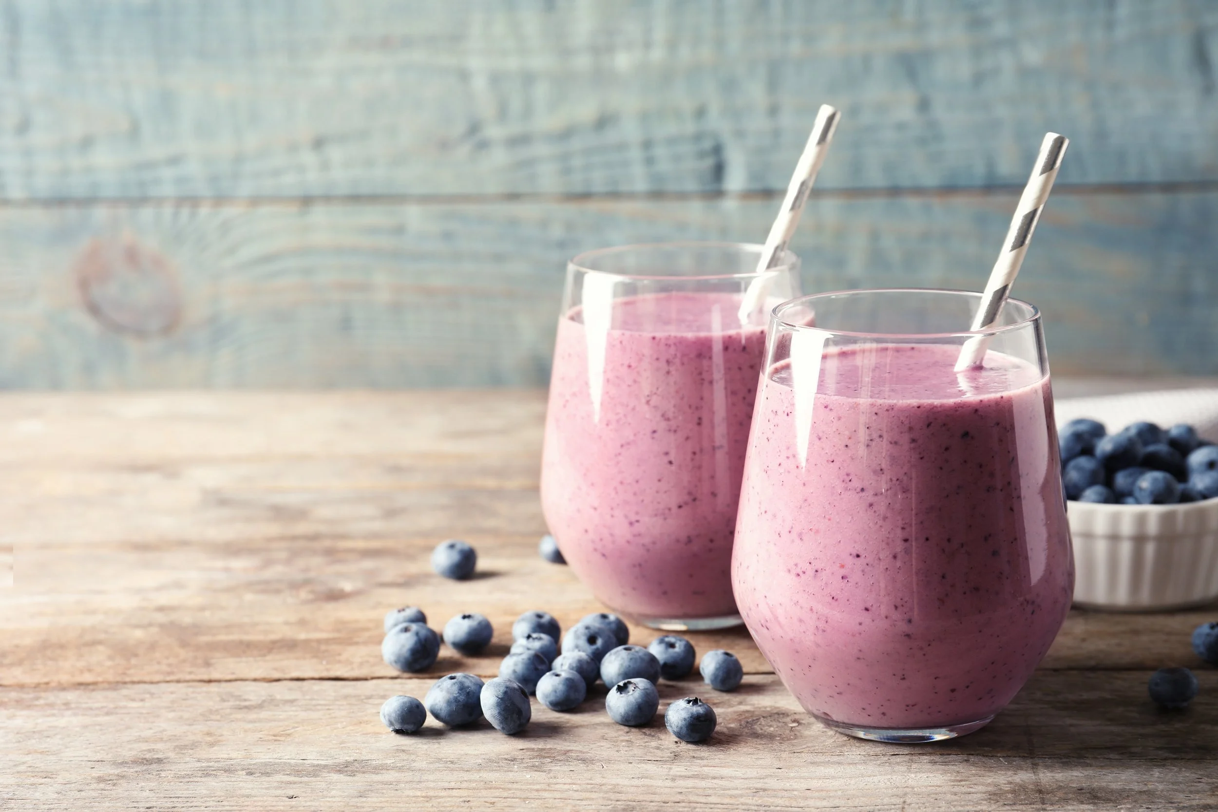 Two glasses of pink blueberry smoothie with striped paper straws on a wooden surface, with scattered blueberries in the foreground and a bowl of blueberries in the background.
