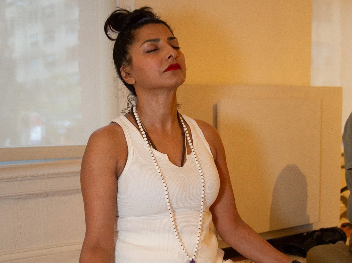 A woman with dark hair tied up, wearing a white sleeveless top, a long pearl necklace, red lipstick, and makeup, appears to be meditating or relaxing with her eyes closed in a room with neutral-colored walls.