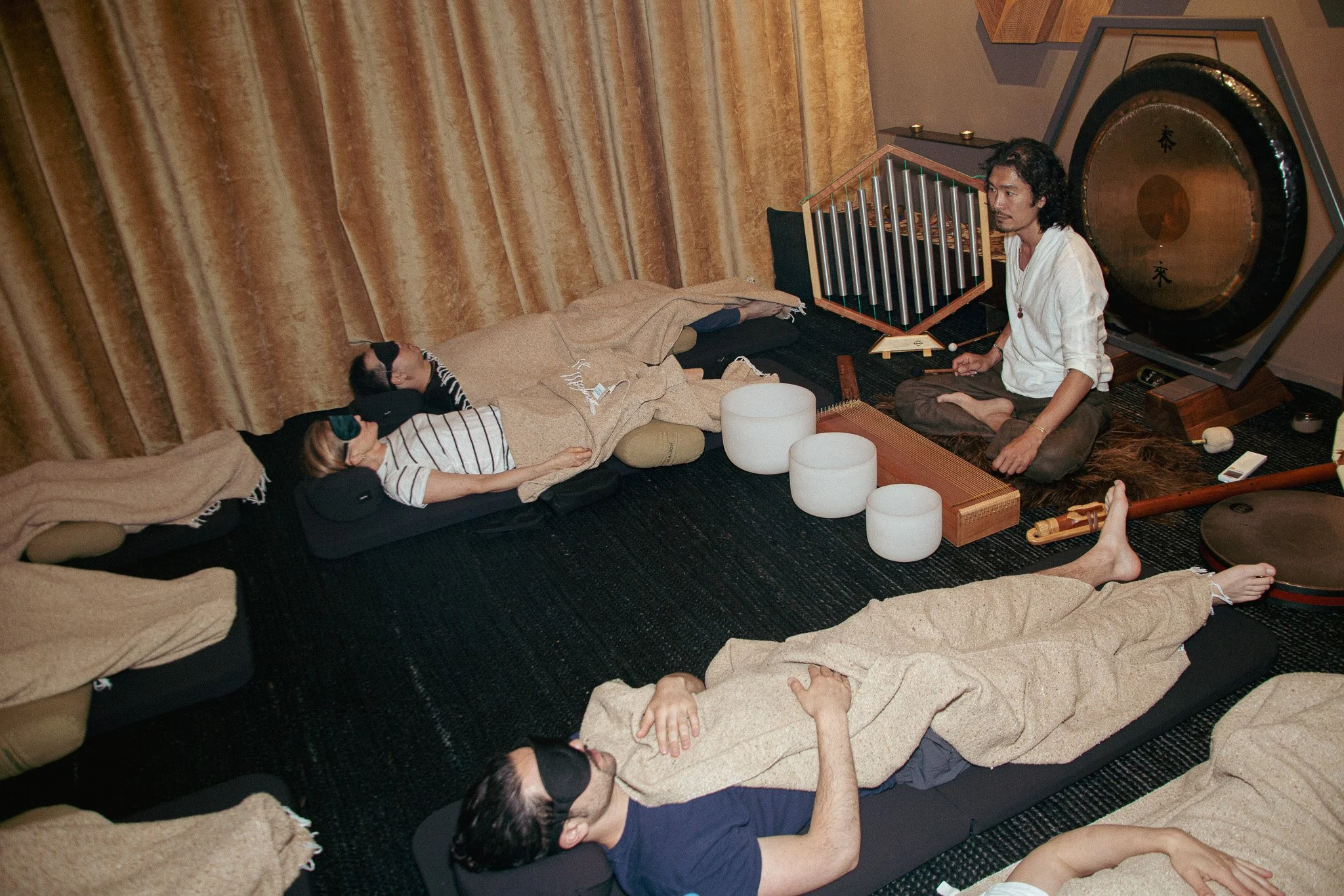 People lying on mats with eyes covered, listening to a man playing singing bowls and a percussion instrument in a meditation or sound healing session in a room with curtains.