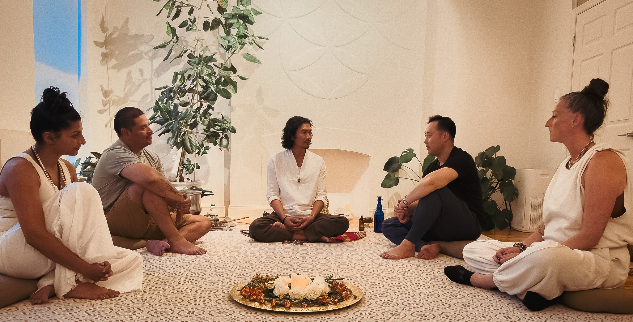 Group of six people sitting cross-legged on the floor in a circle, participating in a meditation or spiritual practice indoors with plants and white walls, one person in the center with eyes closed, others listening or meditating.