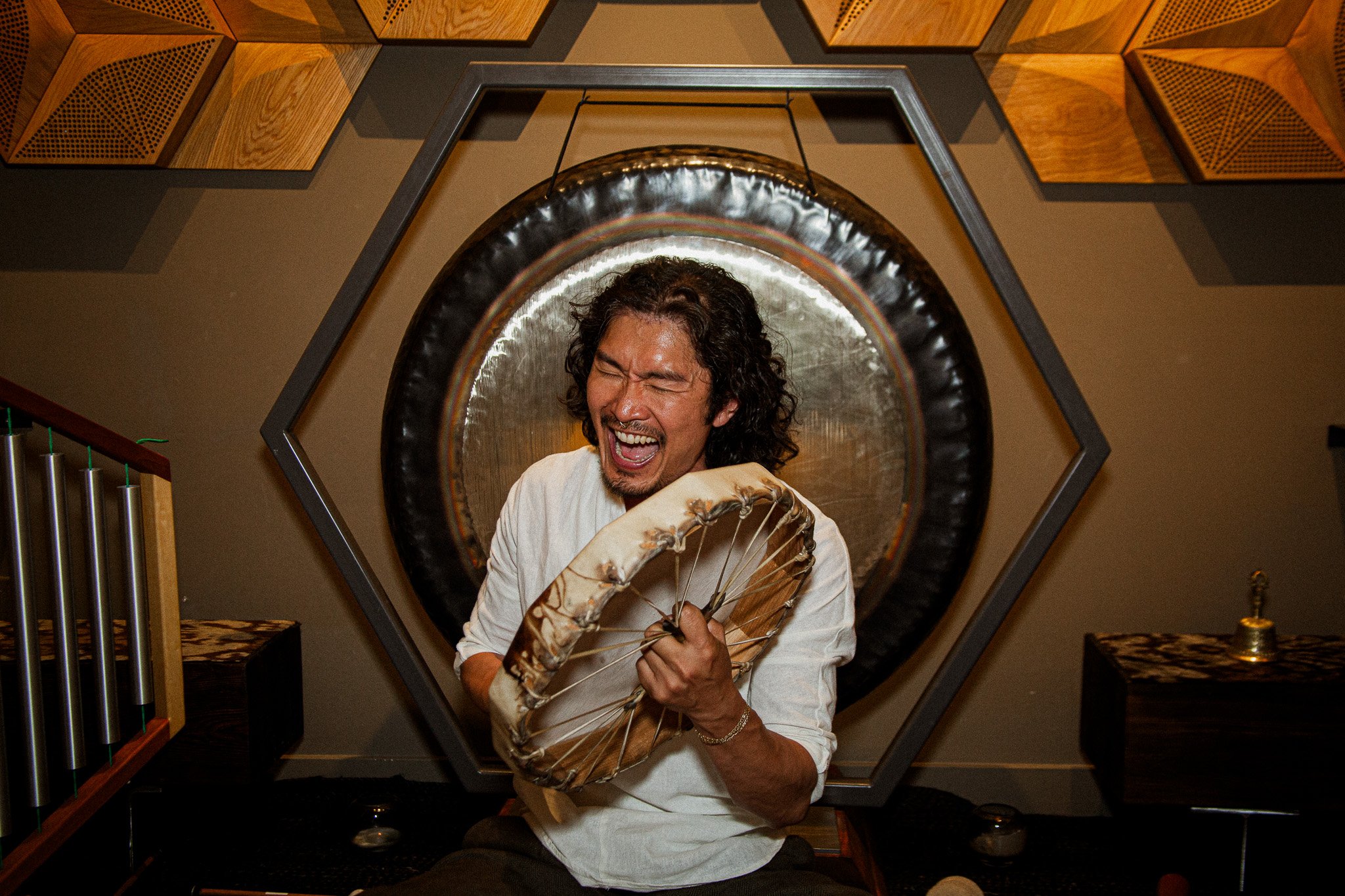 A man with curly hair and a beard, wearing a white shirt, is smiling and playing a tambourine in a music studio. The background features acoustic panels and a gong.