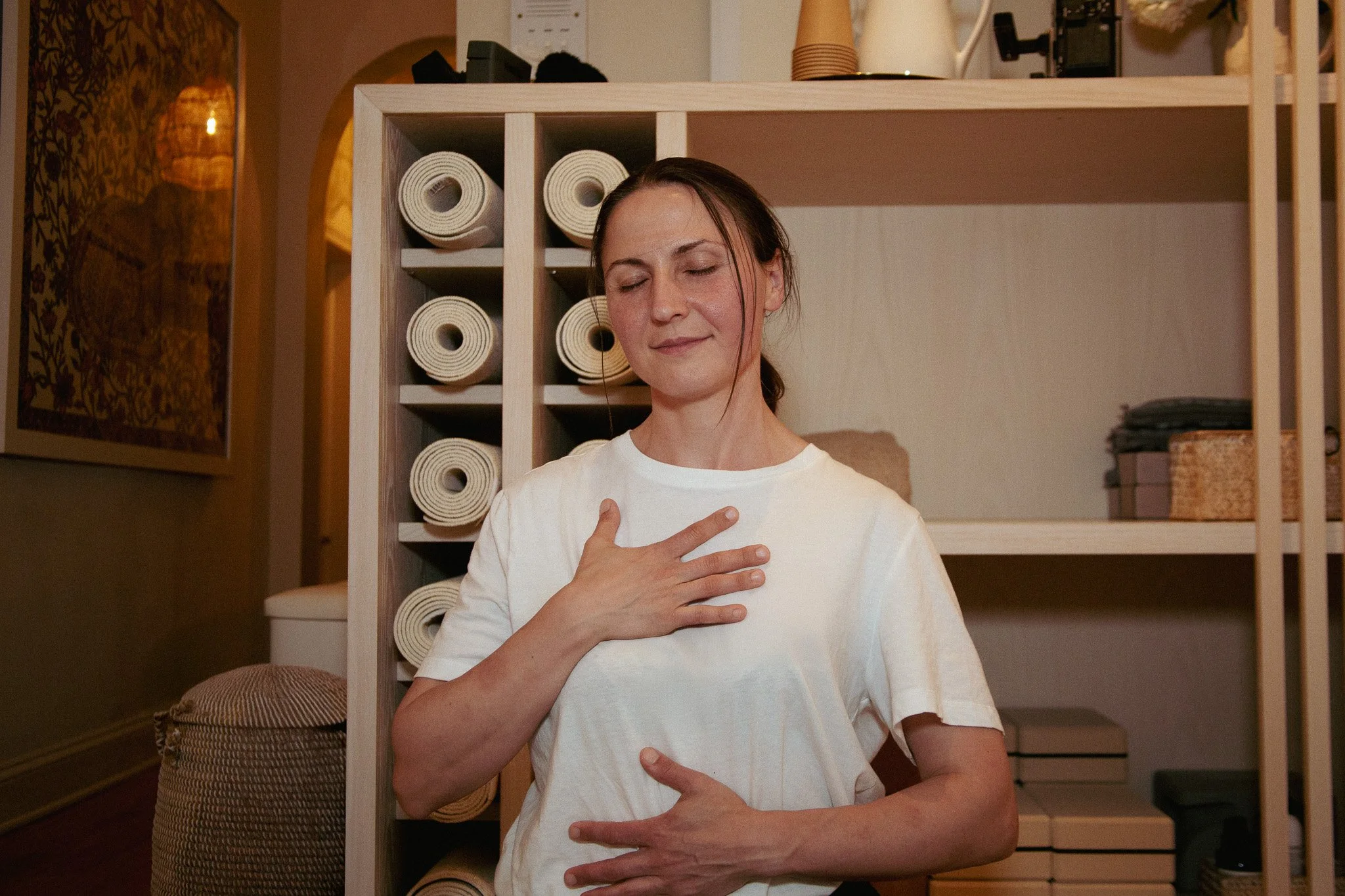 A woman with brown hair wearing a white shirt, standing indoors with her eyes closed, touching her chest and stomach with both hands in a peaceful pose. Behind her is a wooden shelf holding rolled-up towels and other items, and the background features warm lighting and decorative furniture.