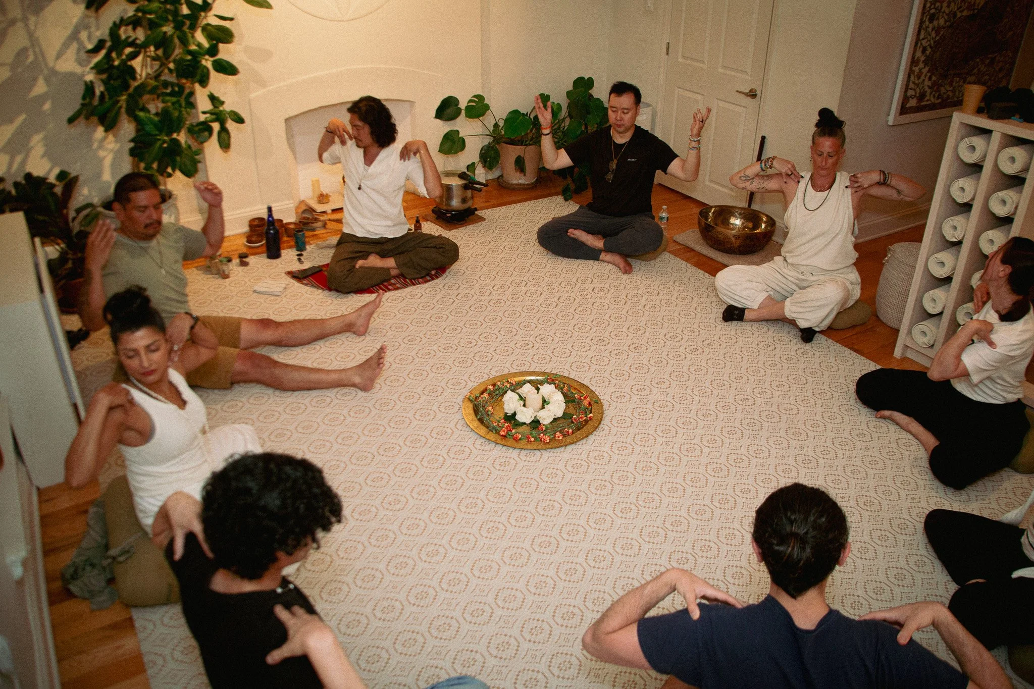 People meditating in a room with a white carpet, a floral centerpiece, and potted plants.