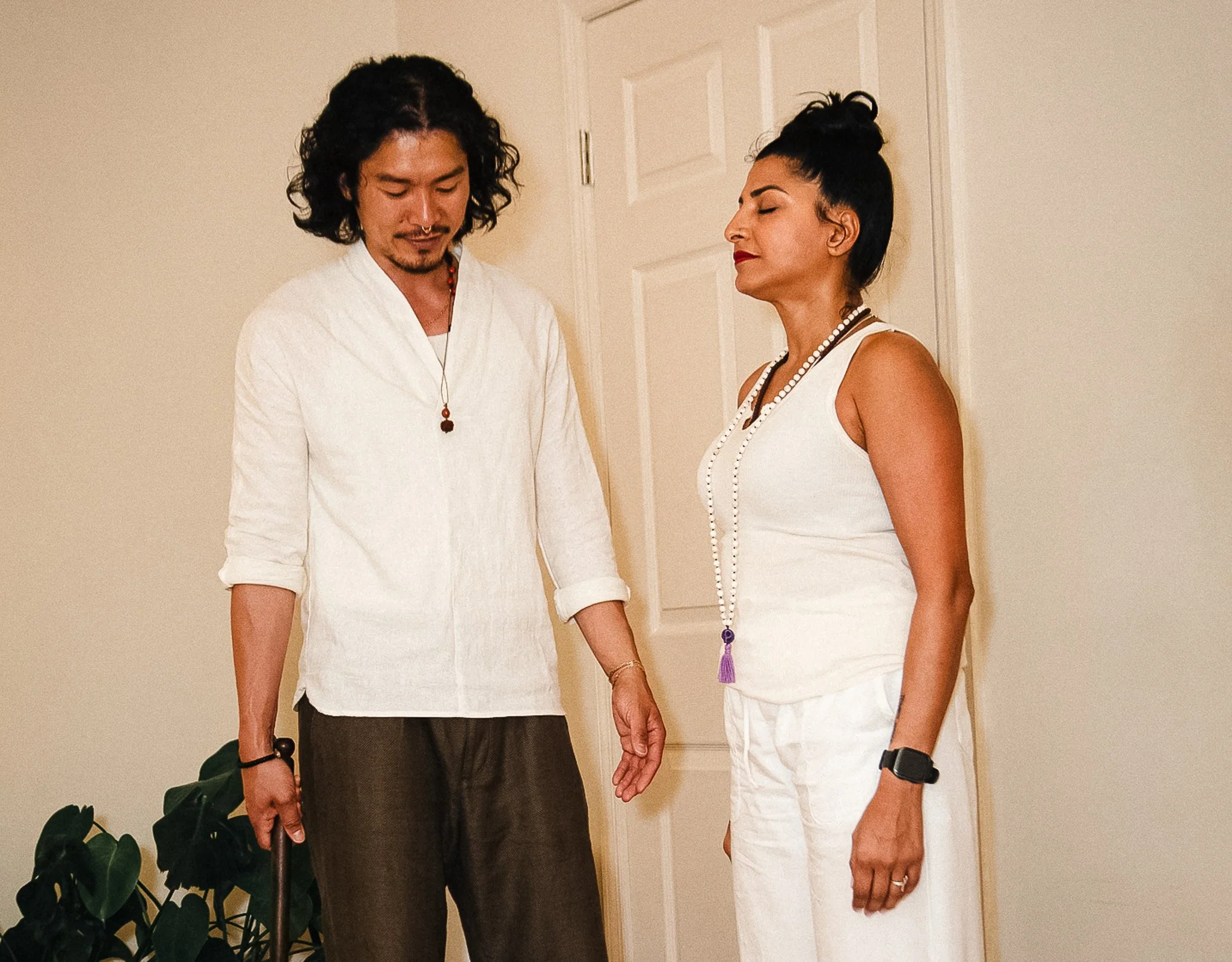 A man and a woman are standing close together, holding hands with their eyes closed in a calm, intimate moment. The man has long, curly dark hair and a light beard, wearing a white shirt and dark pants. The woman has styled dark hair in an updo, wearing a sleeveless white top and white pants. They are standing in a room with light-colored walls and a closed door in the background, next to a leafy houseplant.