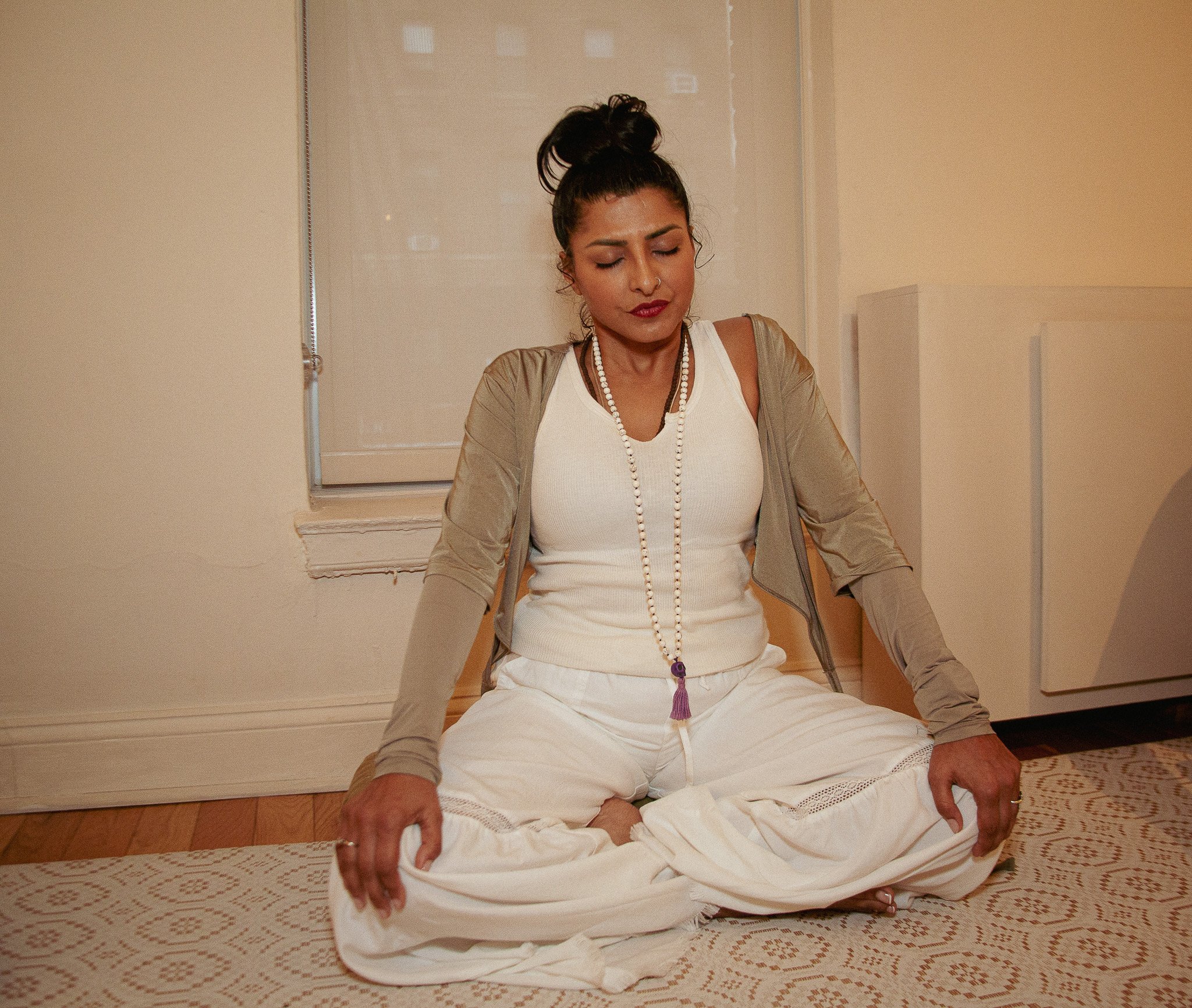 A woman with dark hair tied in a bun, wearing a nose ring, white top, beige jacket, and white pants, sitting cross-legged on a rug with her eyes closed and hands resting on her knees in a meditative pose indoors.