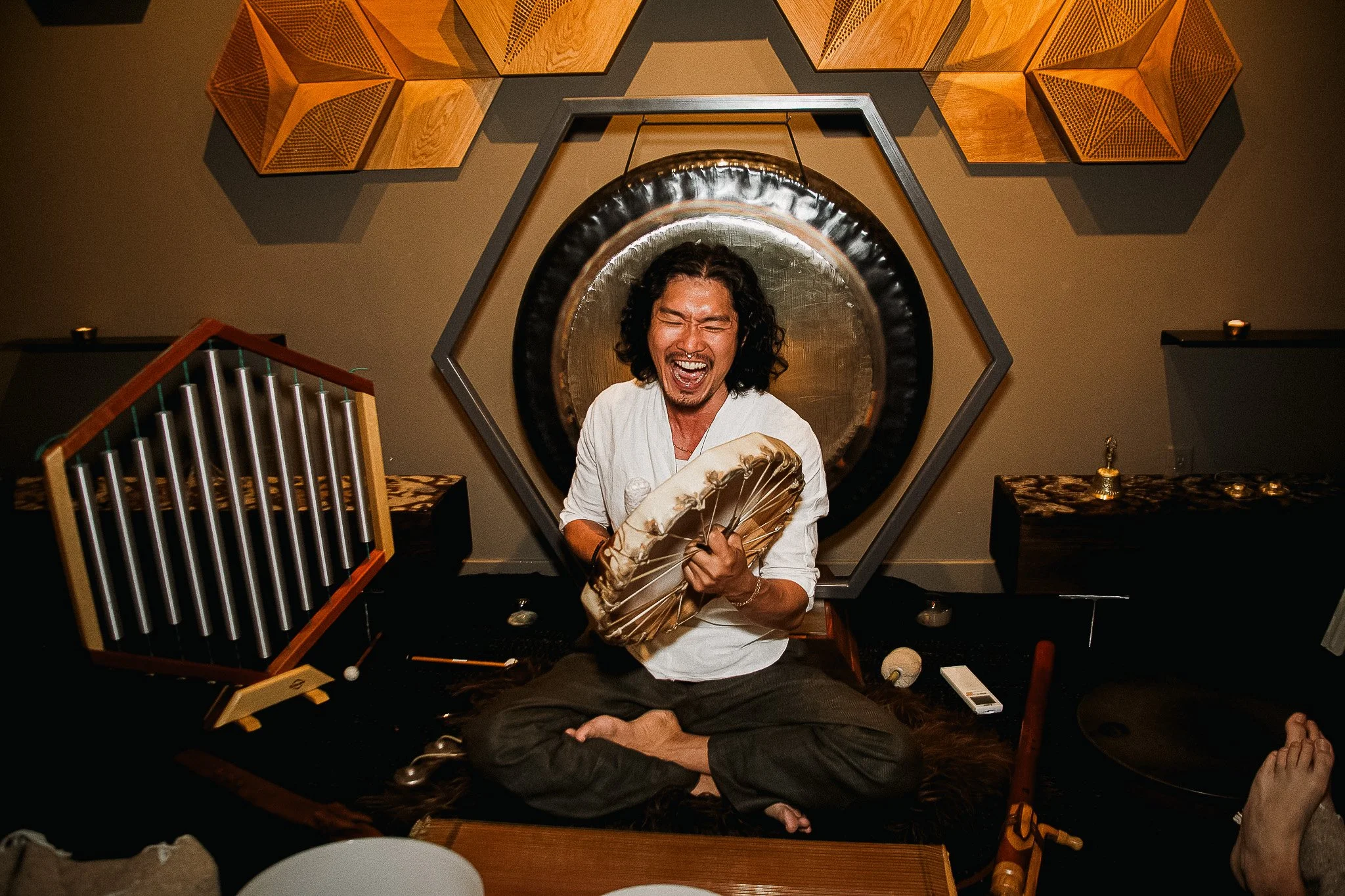 A man with long curly hair, wearing a white shirt, is standing in front of a gong and smiling while holding a drumstick in his right hand. The background has geometric wooden wall art and sound equipment.