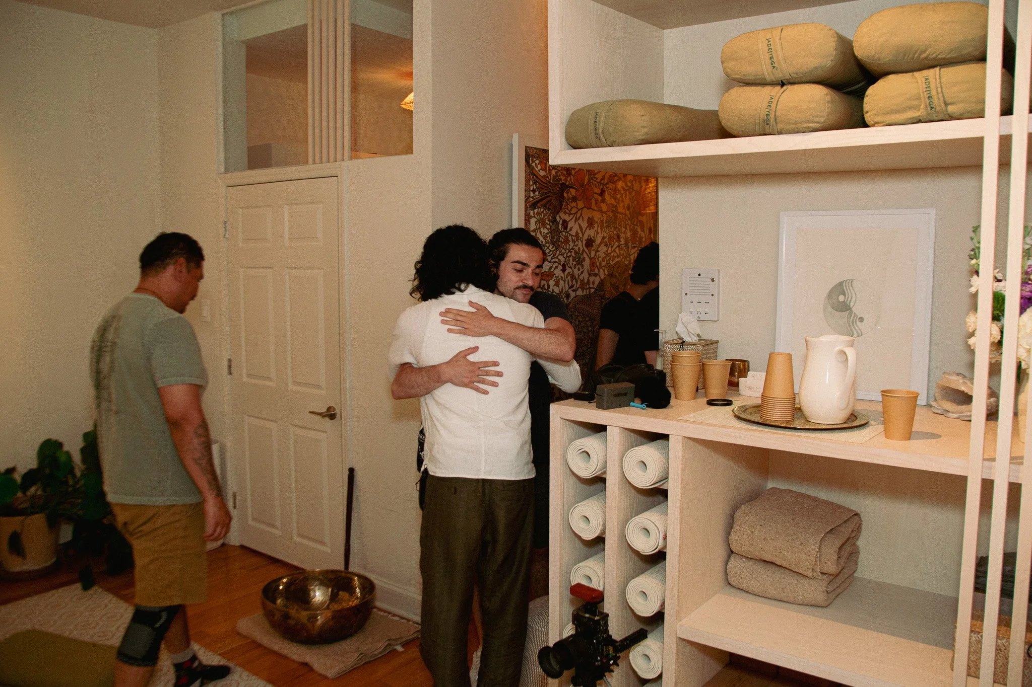 A group of people gathered in a cozy, well-lit room, hugging each other near a wooden shelf filled with rolled towels, pottery, and decorative items, with some individuals smiling and embracing.