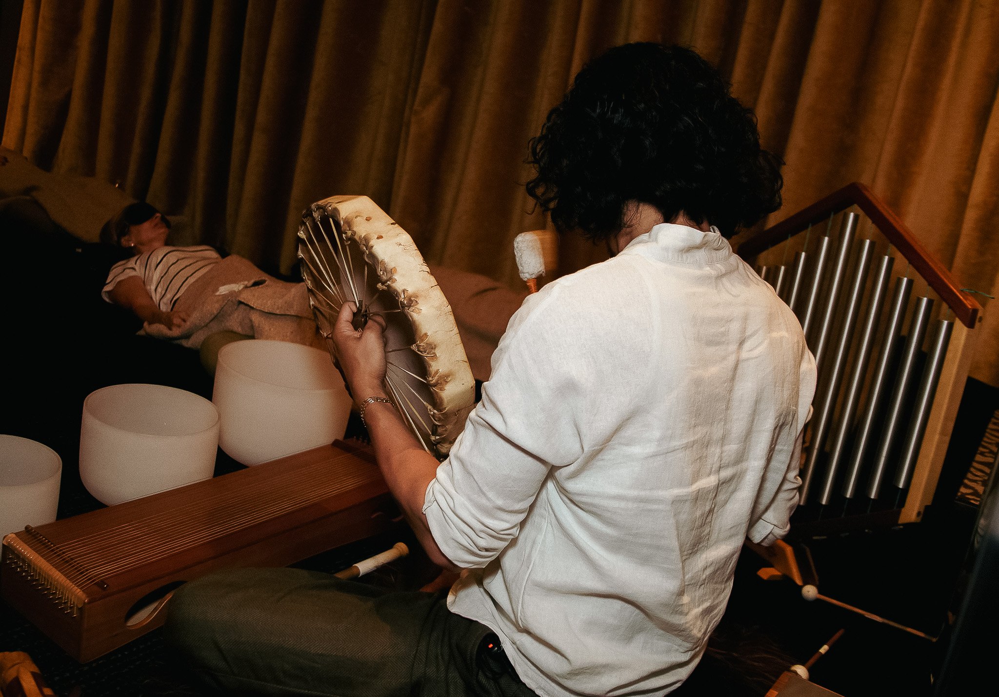 A woman with curly dark hair playing a hammered dulcimer, viewed from behind, in a room with brown curtains and a man lying on a couch in the background.