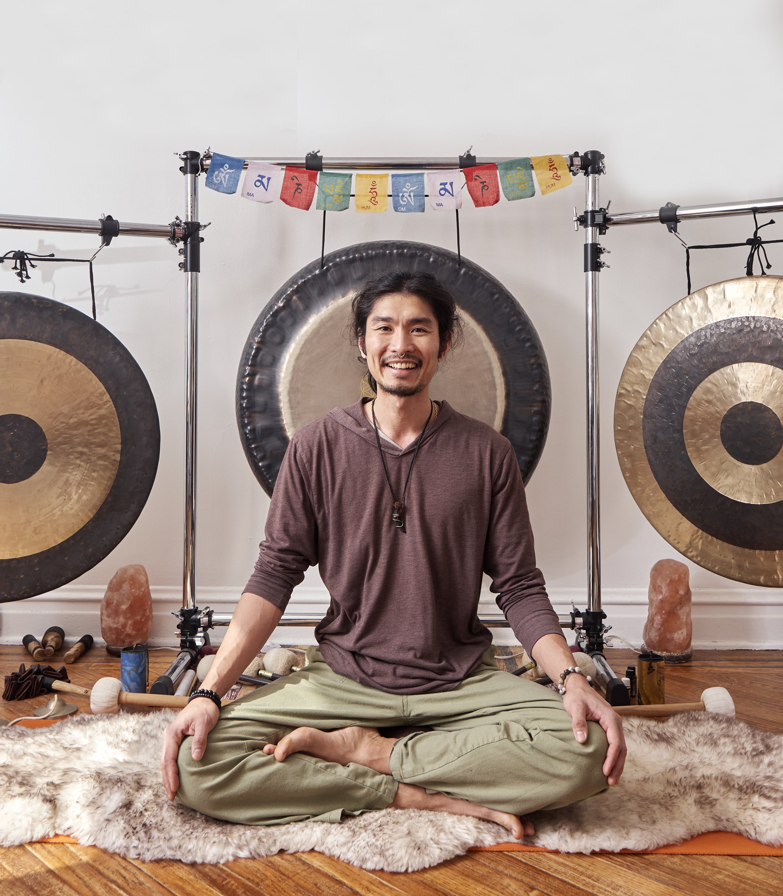A man practicing meditation on a fluffy rug surrounded by musical instruments and gongs, with Tibetan prayer flags hanging above his head, in a serene indoor setting.