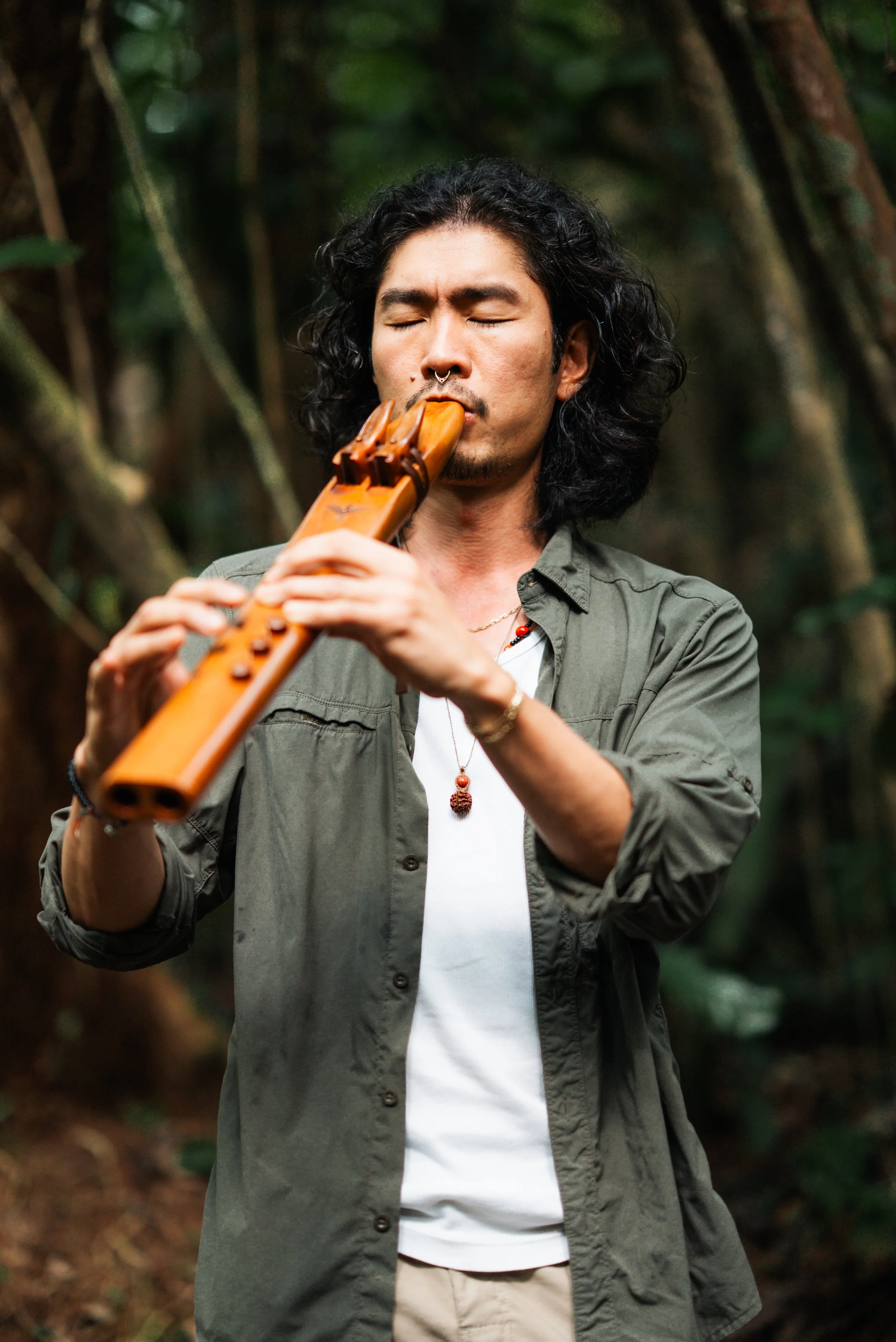Man with long black curly hair and closed eyes playing a wooden flute in a forest.
