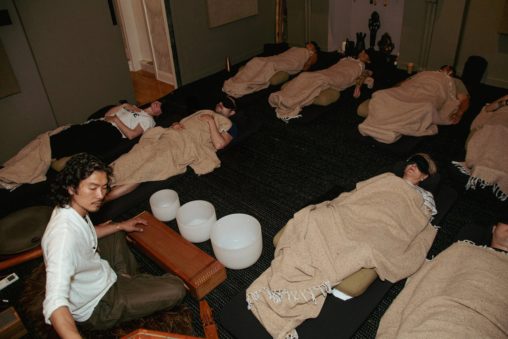 Several people are lying on cushions on the floor, some with sleep masks, appearing to sleep or rest, with a person playing a musical instrument nearby.